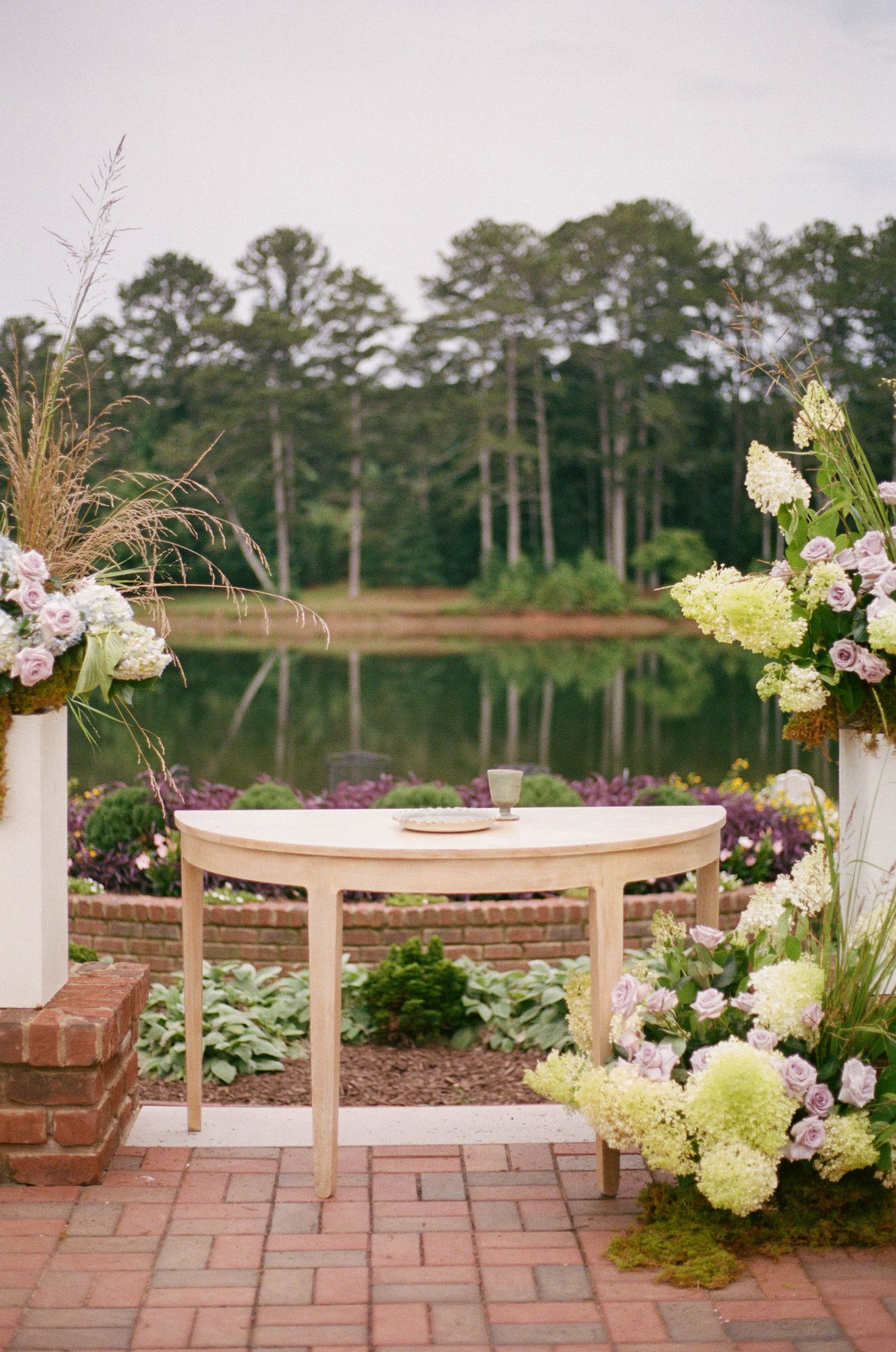 Elegant outdoor wedding ceremony altar with hydrangea floral arrangements flanking a white table overlooking a serene lake.