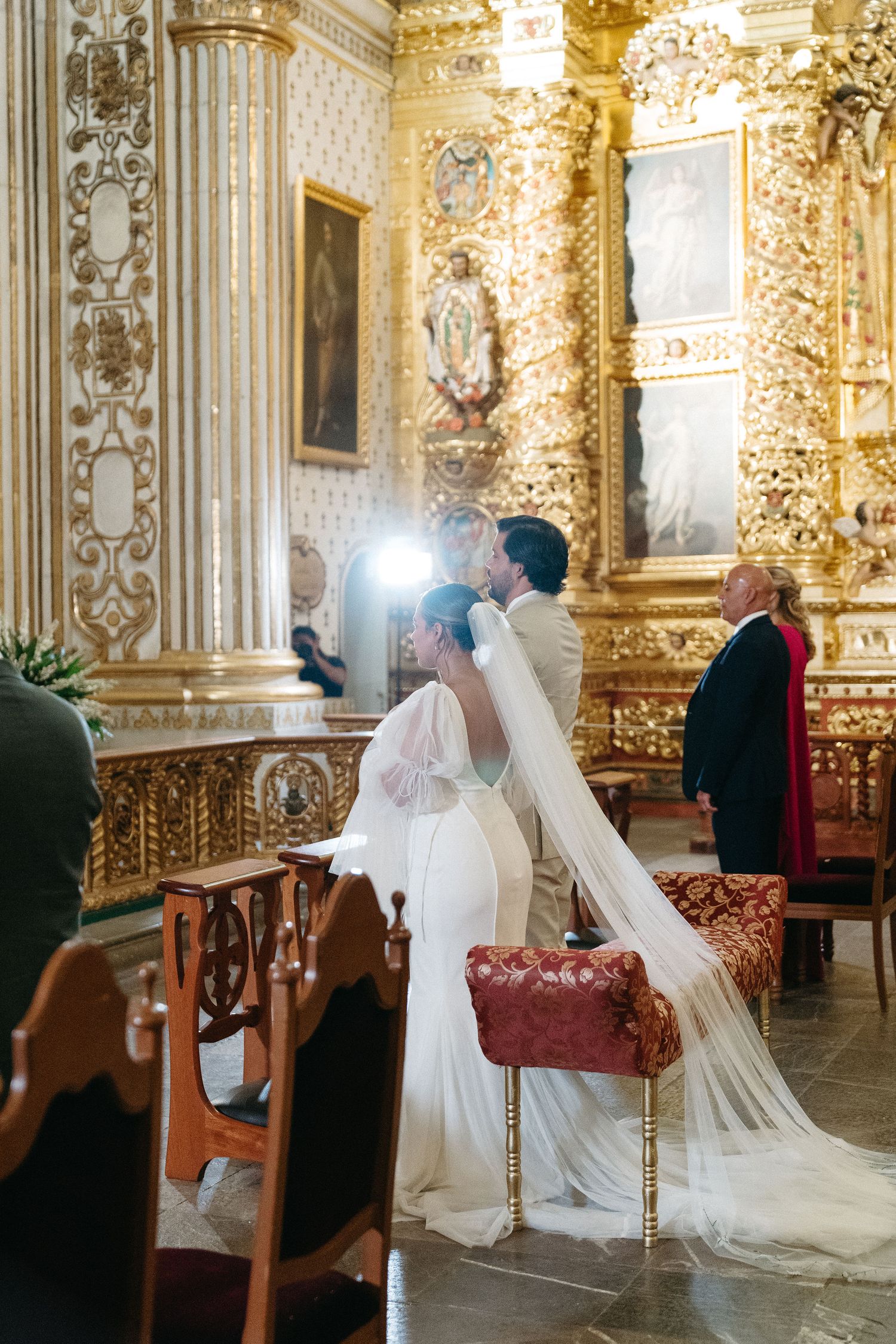 A wedding ceremony in an ornate golden baroque church interior with flowing white dress and veil.