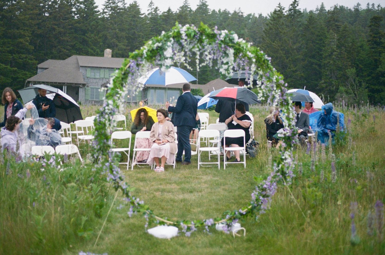 An outdoor wedding reception setup with white chairs and floral arch in a scenic field.