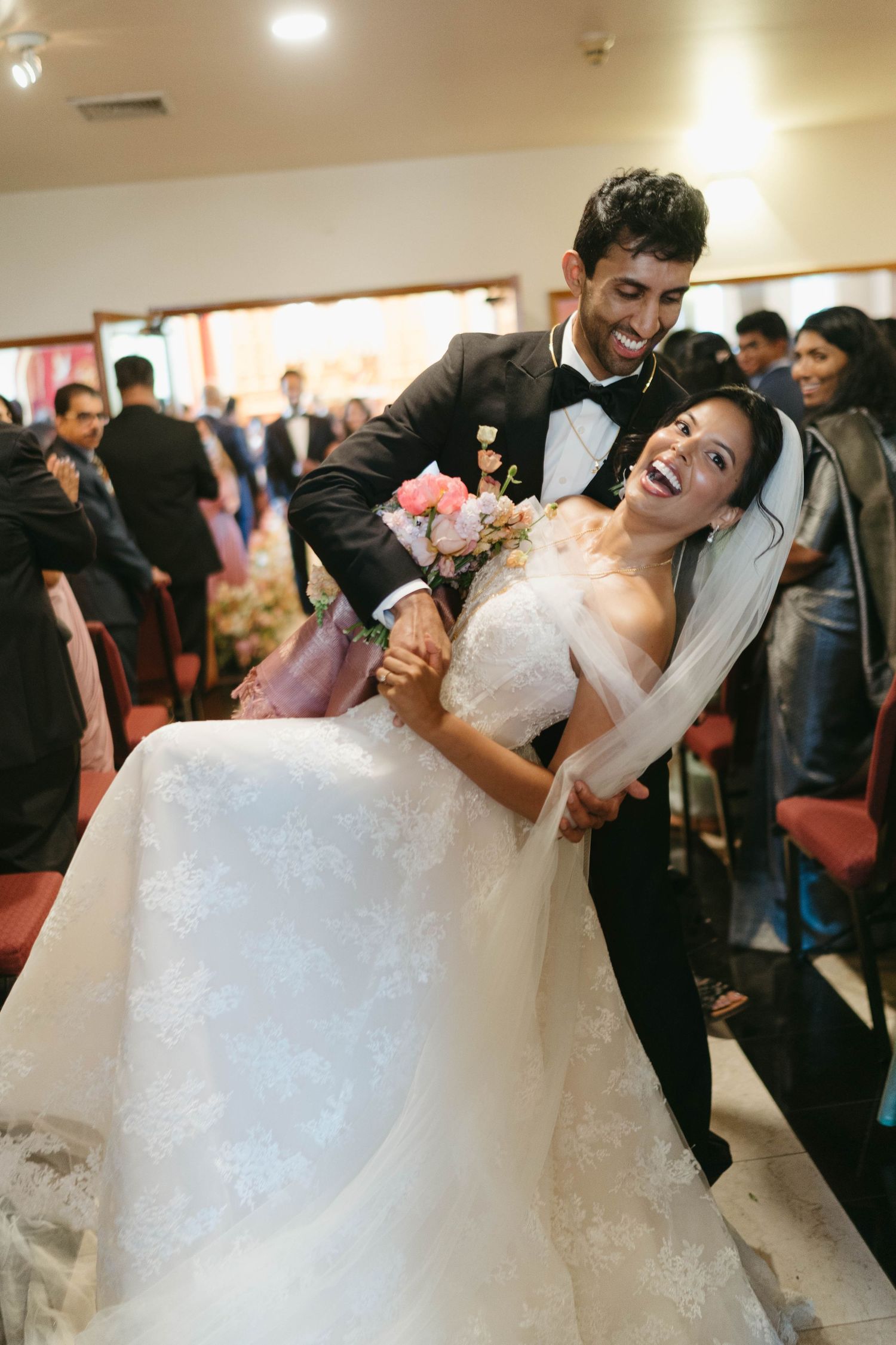 Joyful bride and groom laughing together during church recessional, bride holding pink bouquet in lace gown.