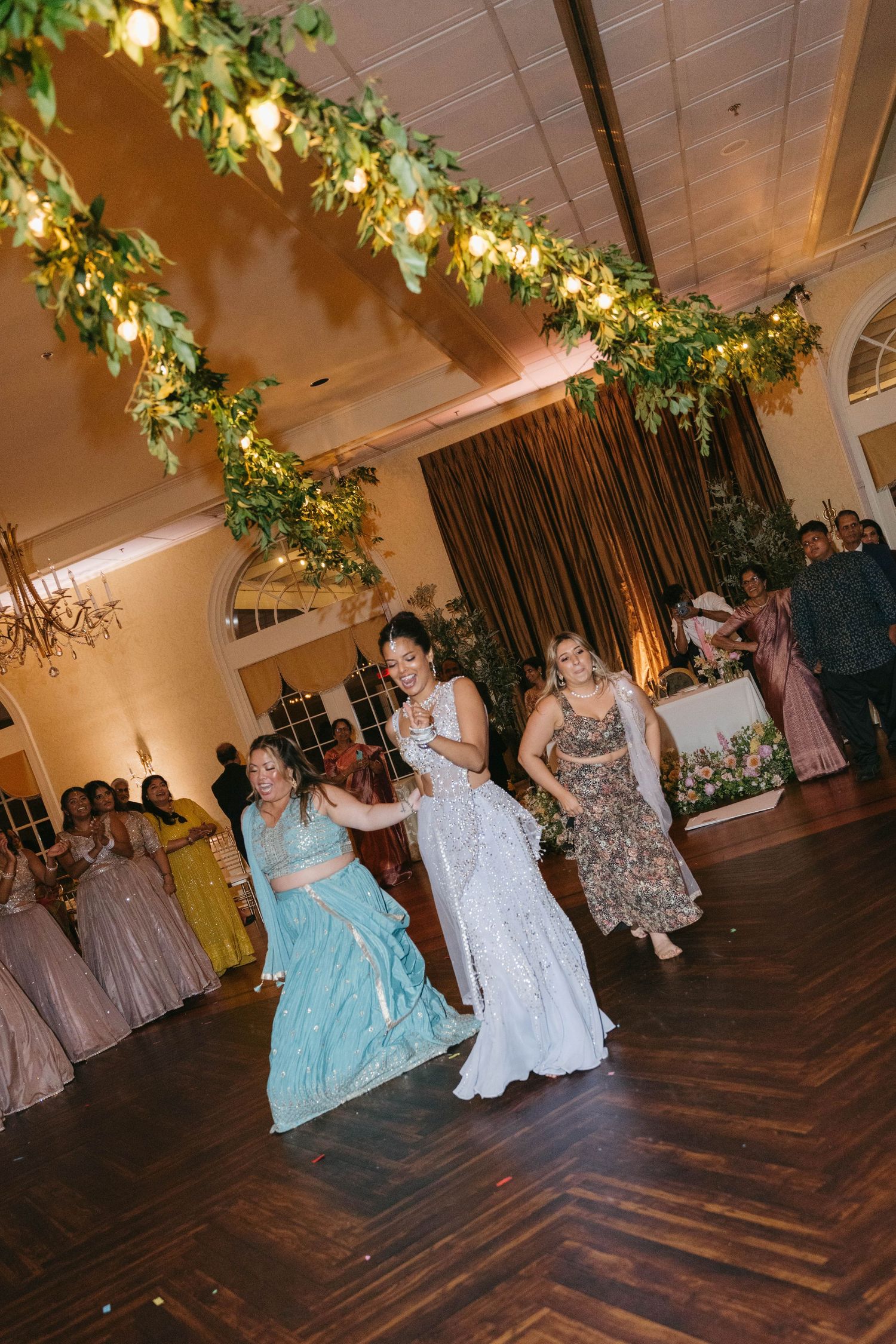Guests dance joyfully on the wooden floor at a wedding reception adorned with lush greenery and warm string lights.