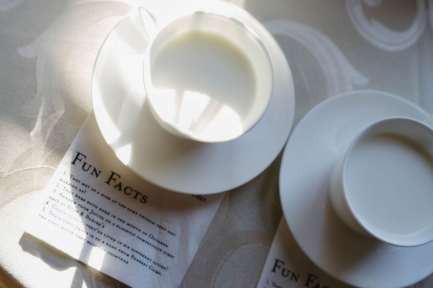 Two white ceramic cups and saucers rest on a Fun Facts paper pamphlet in soft natural light.
