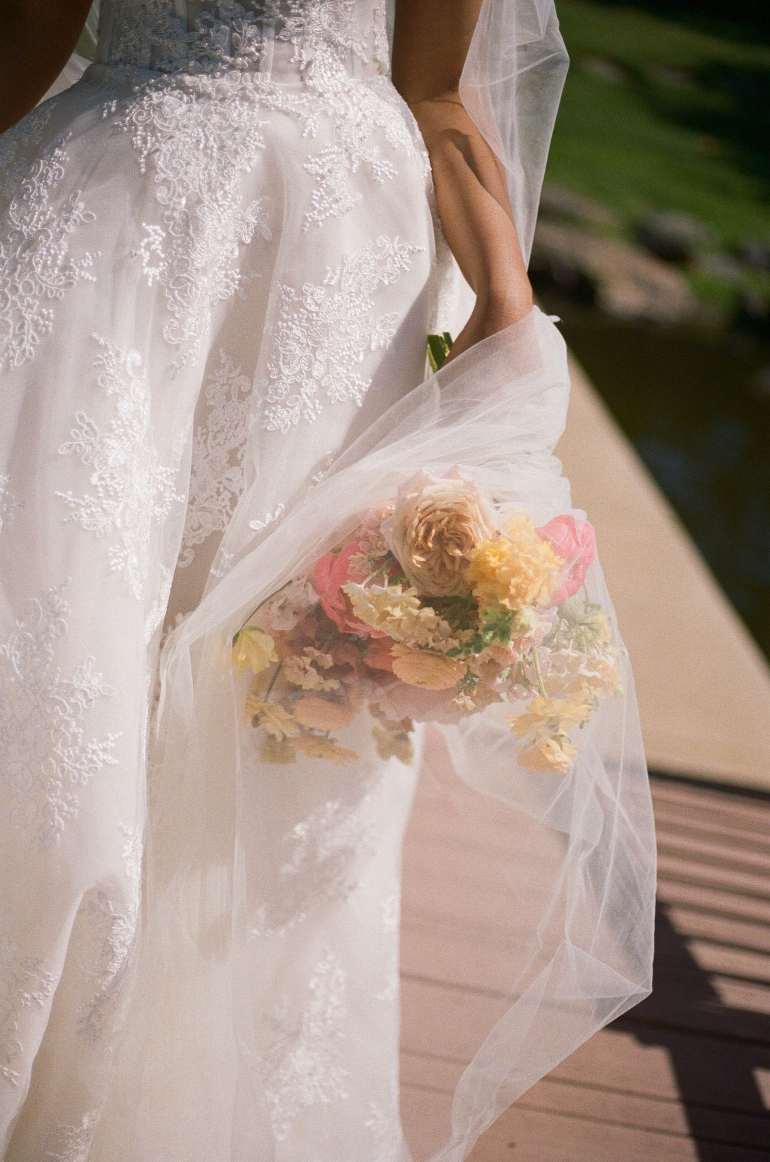 Close-up of brides lace wedding gown and veil with colorful pink and yellow bouquet outdoors.