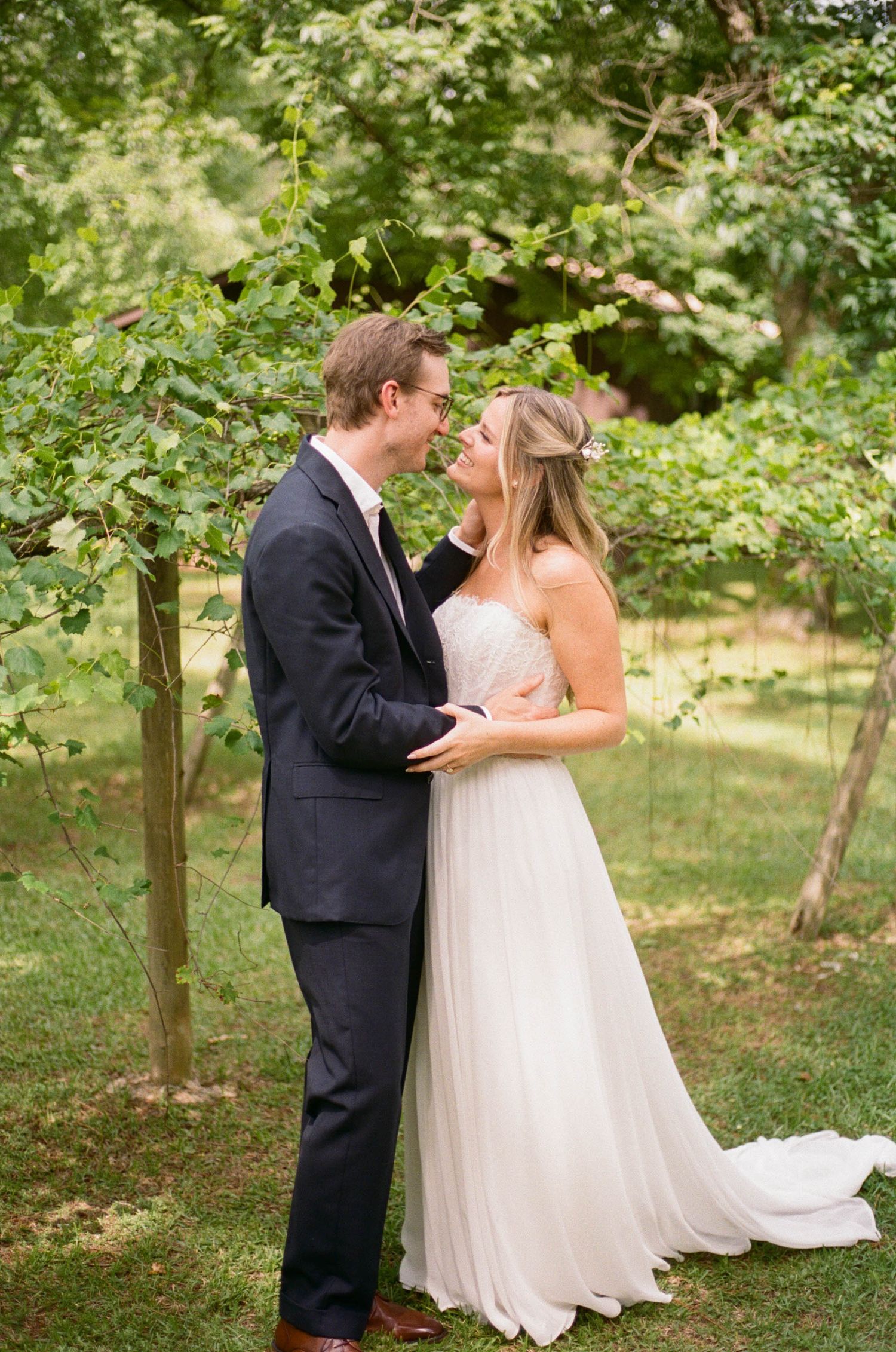 Happy bride and groom embracing under leafy trees in a lush green outdoor wedding portrait.