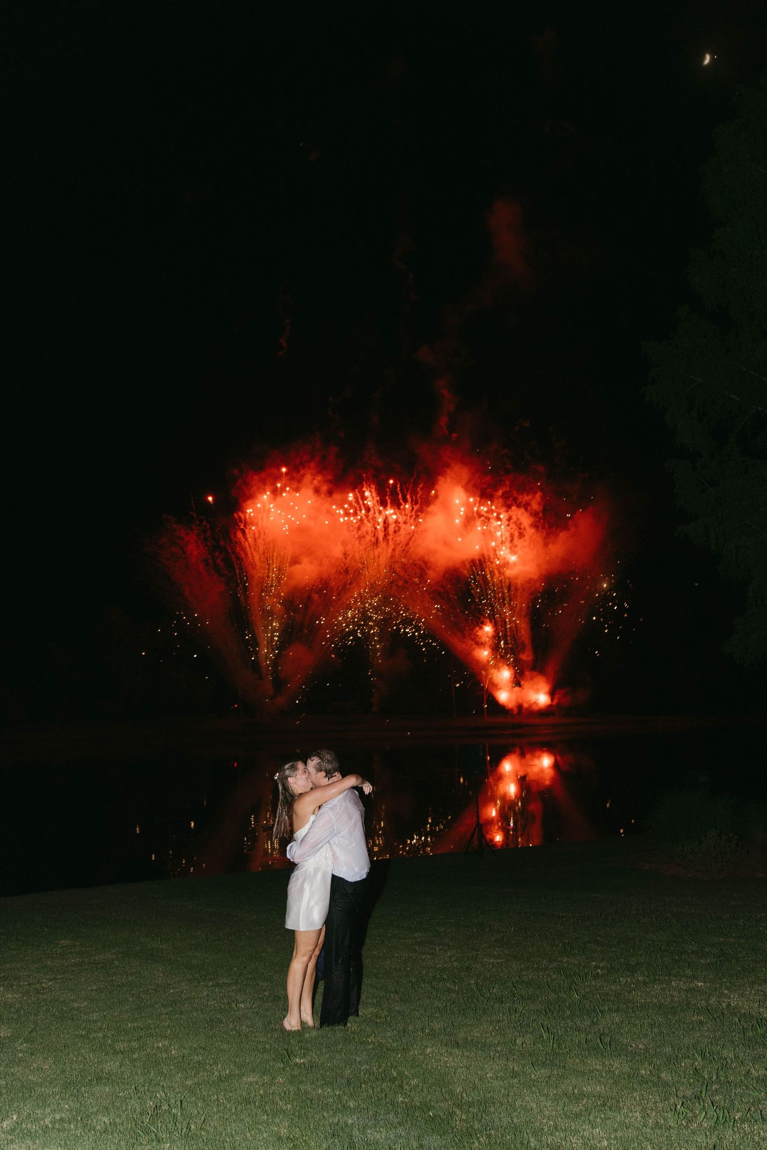 Couple kissing on lawn with stunning red fireworks reflecting over a lake at night.