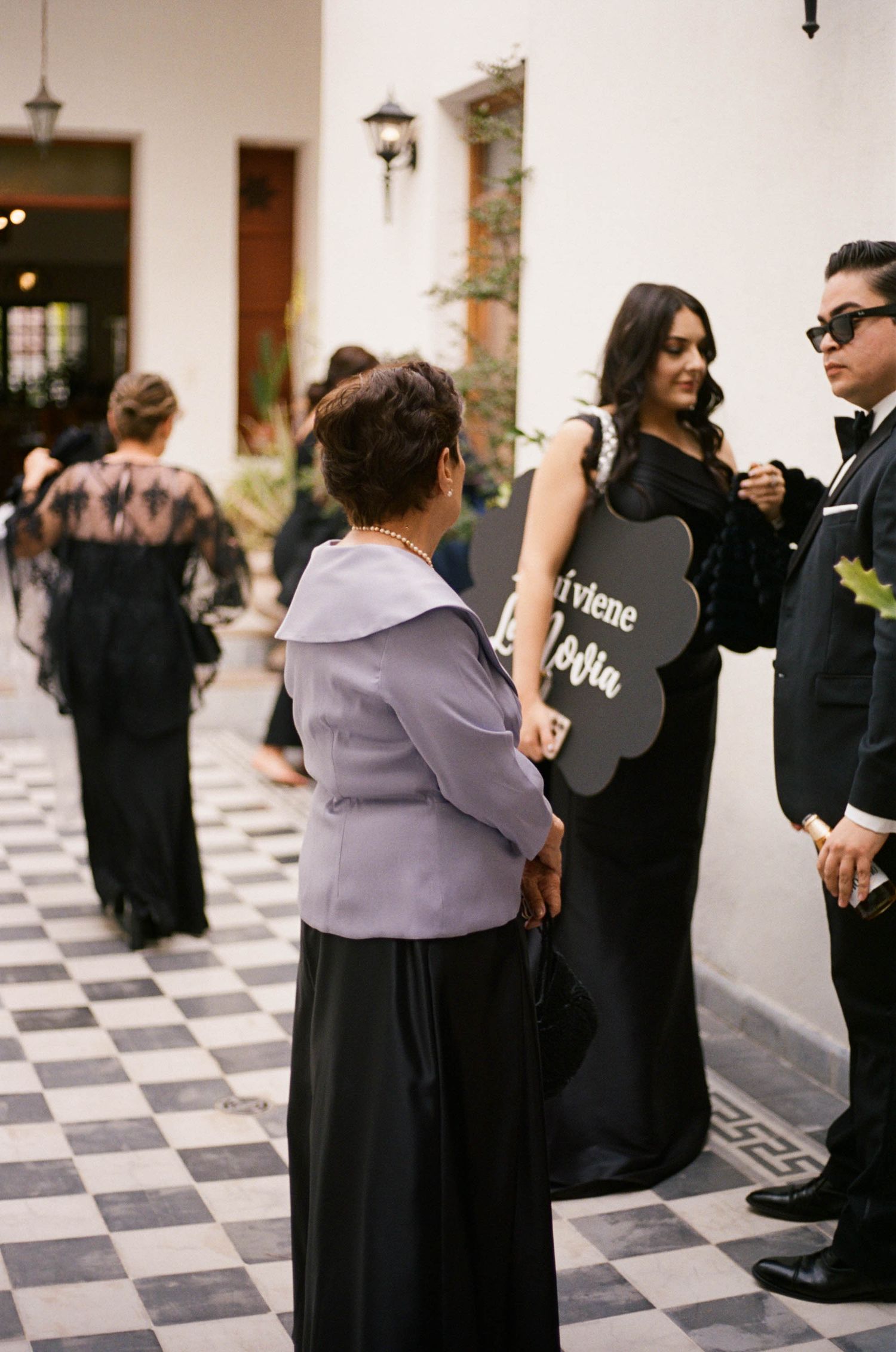 People gathered in formal attire along a hallway with classic black and white checkered flooring.