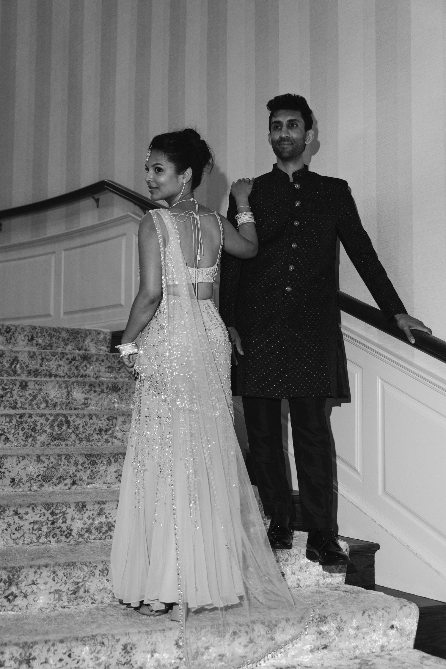 Elegant couple in Indian wedding attire poses on grand staircase, woman in beaded gown, man in dark sherwani.