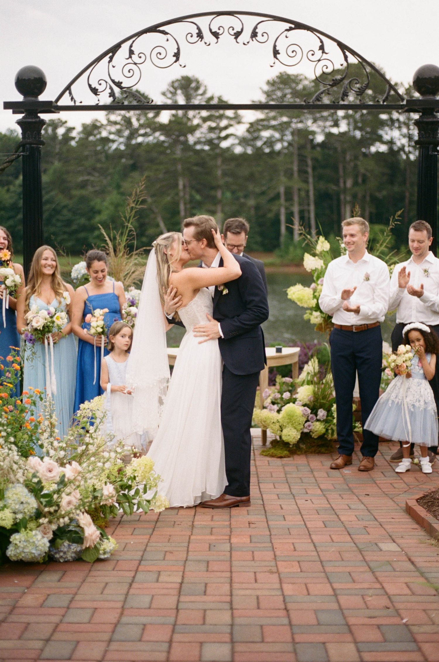 Newlyweds share first kiss under ornate iron arch surrounded by wedding party and lush floral arrangements.