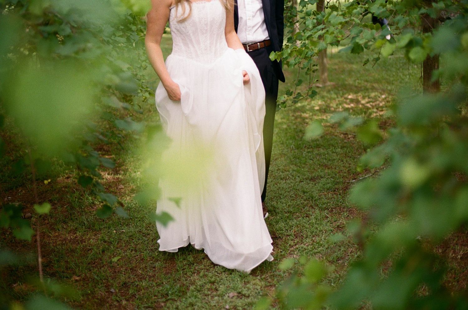 Bride in flowing white strapless gown stands with groom among lush green garden foliage.