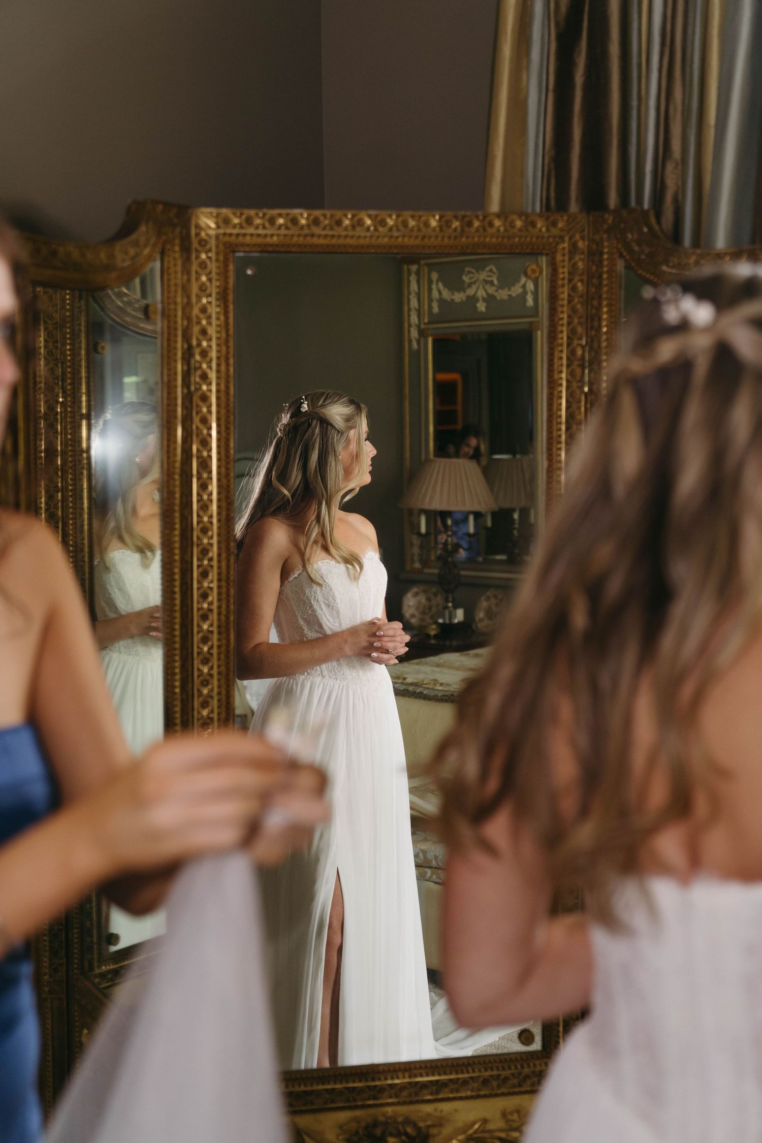 Bride in white gown with floral crown reflected in ornate gold mirror during wedding prep.