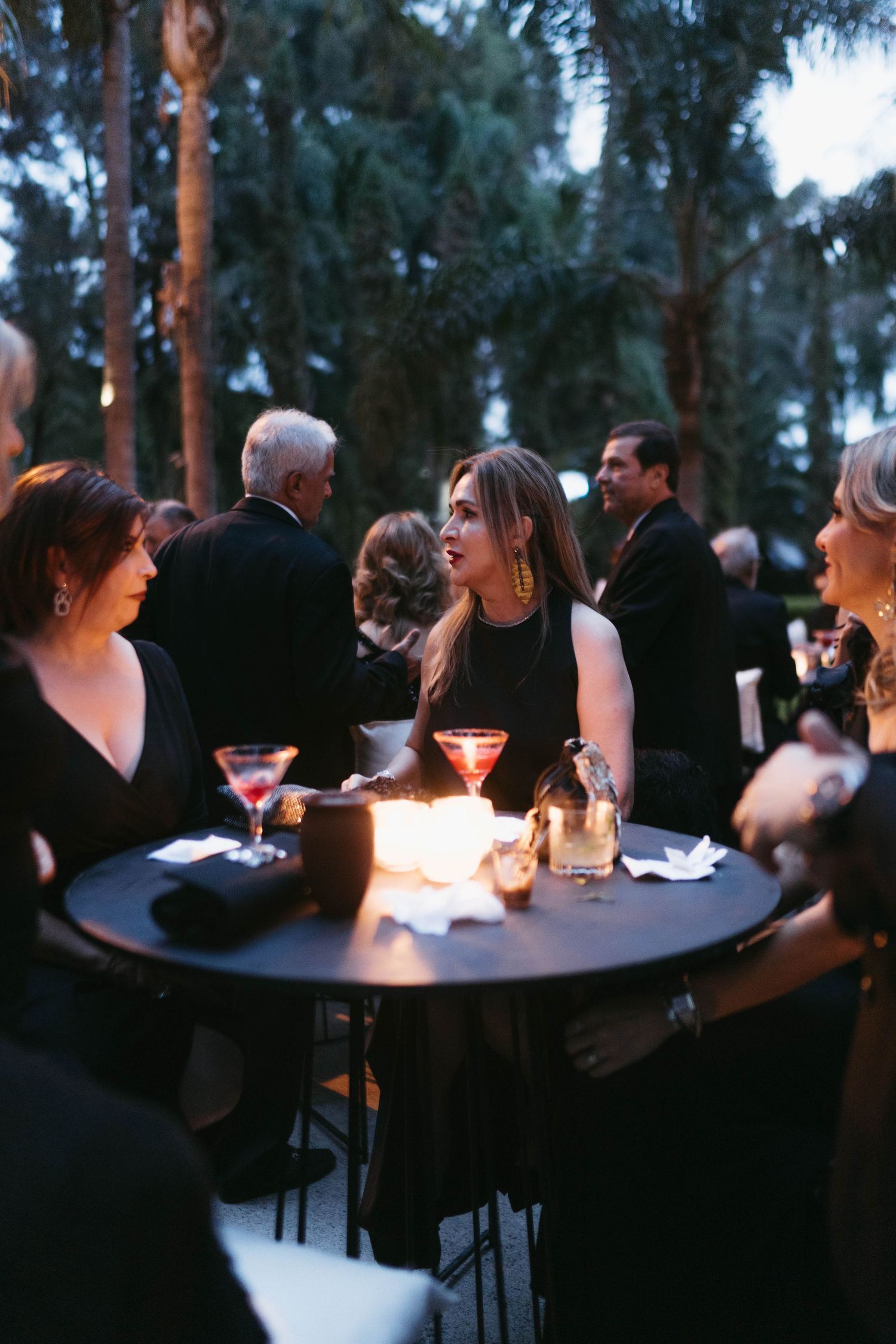 Evening outdoor reception with guests seated at round tables under trees.