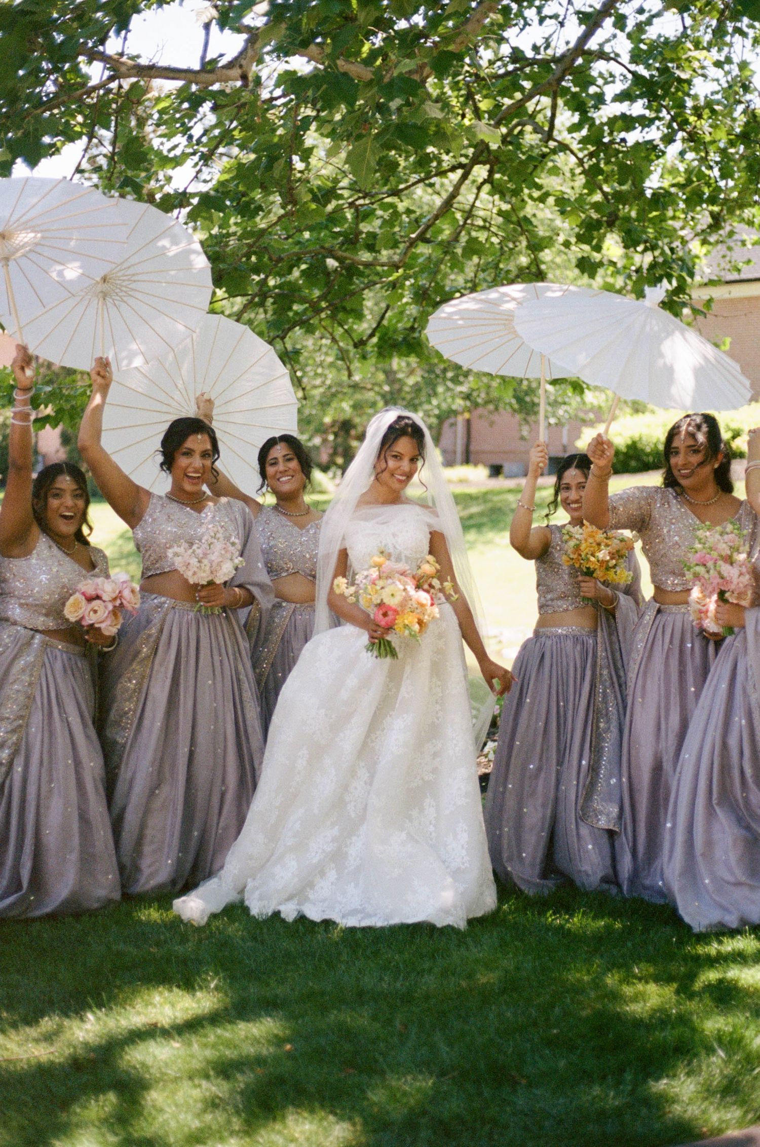 Bride in white lace gown with bridesmaids in lavender lehengas holding white parasols outdoors.