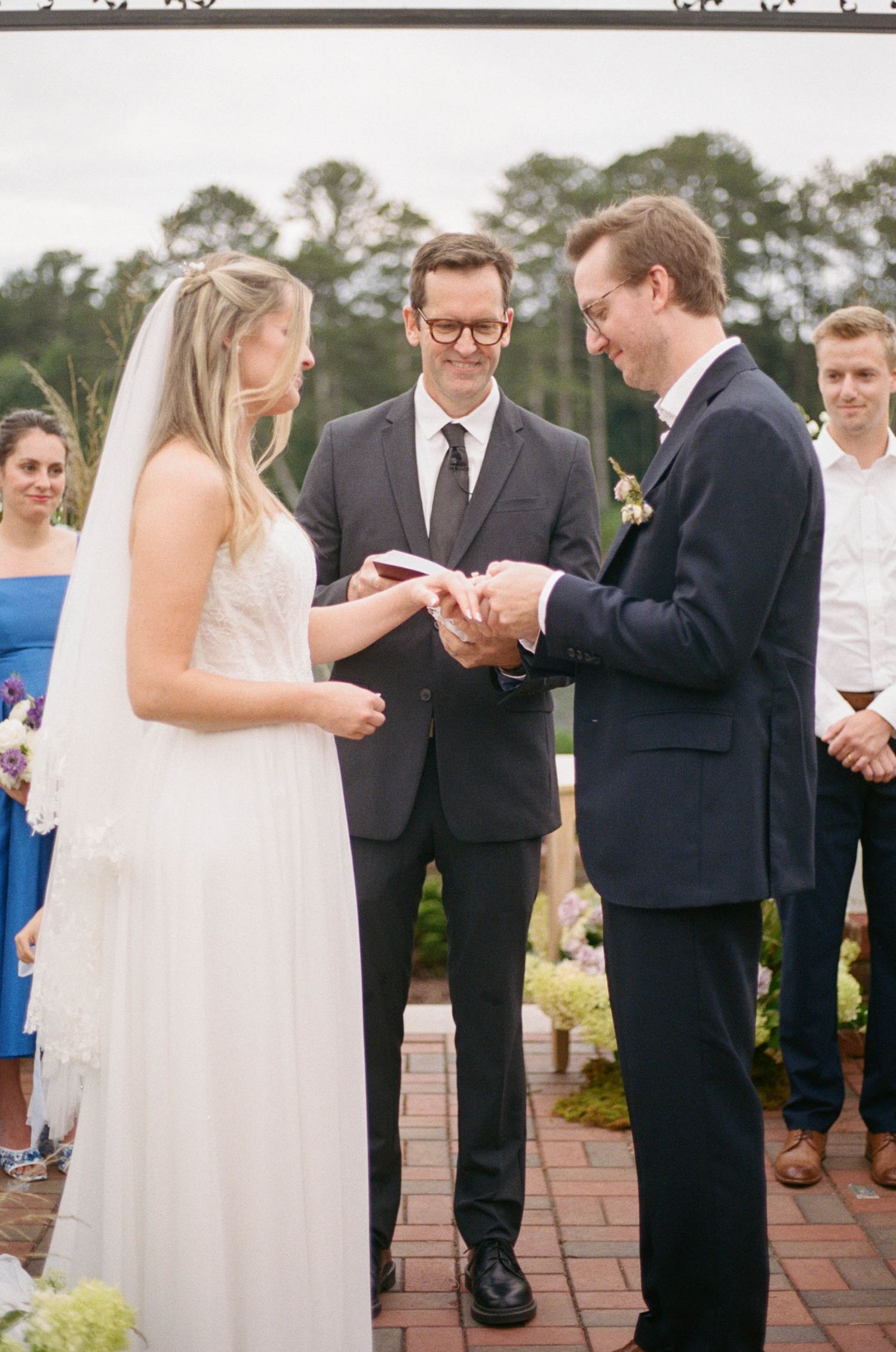 Bride and groom exchange rings during outdoor wedding ceremony with officiant smiling in background.