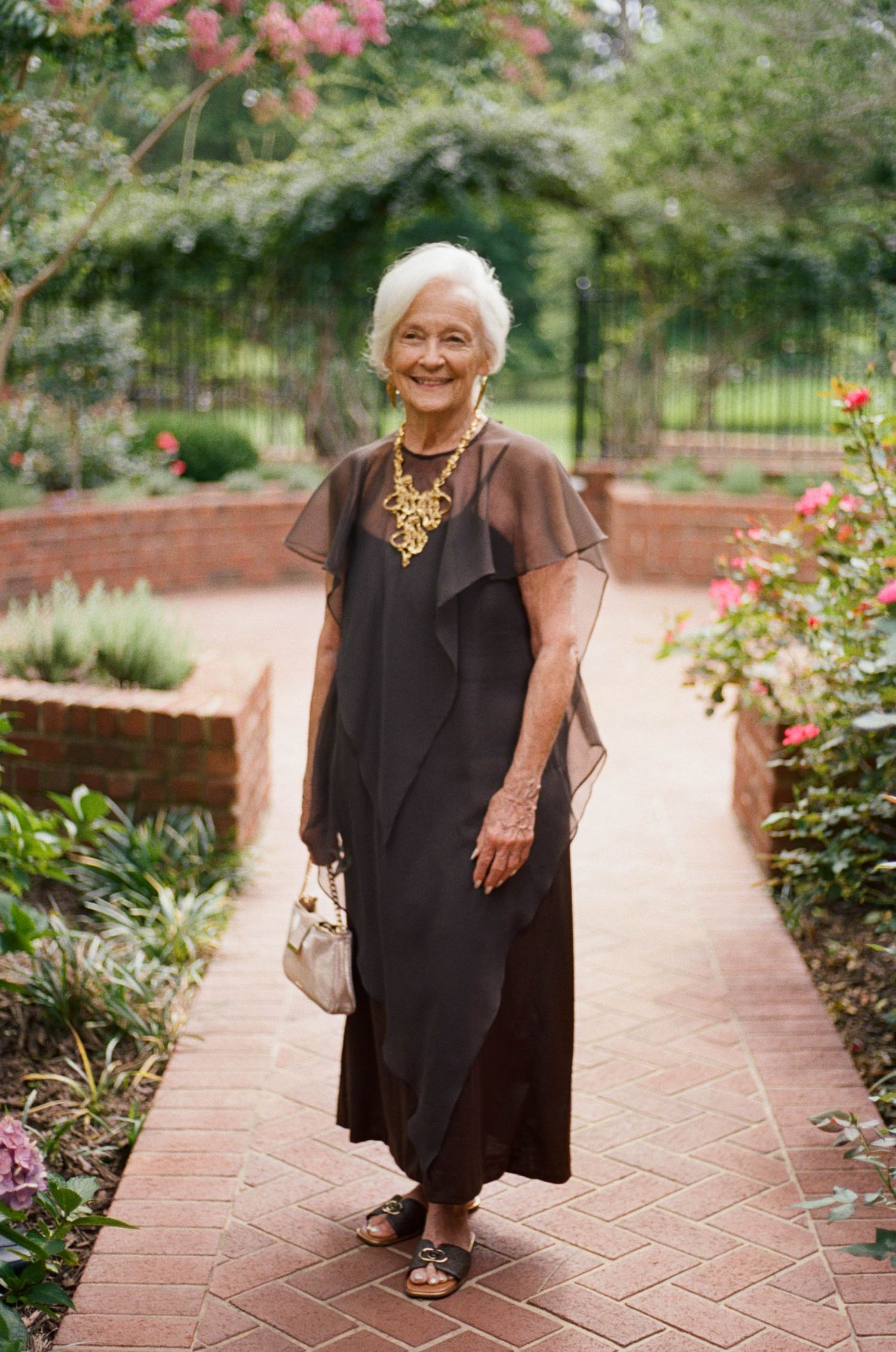 Elegant elderly woman in black dress with gold necklace walks garden brick pathway.
