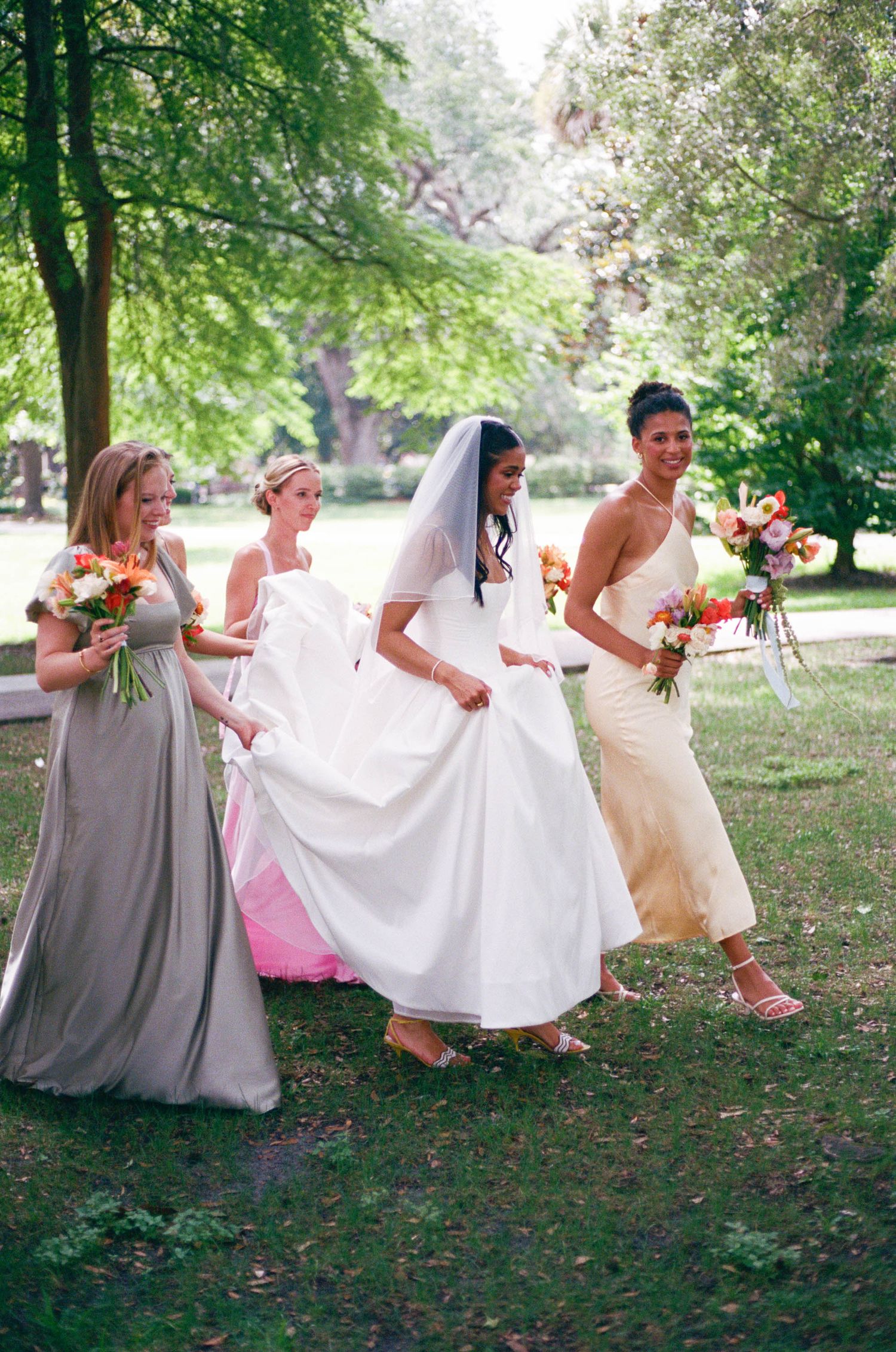 Bridesmaids in pink and gray dresses pose with bouquets under green trees.