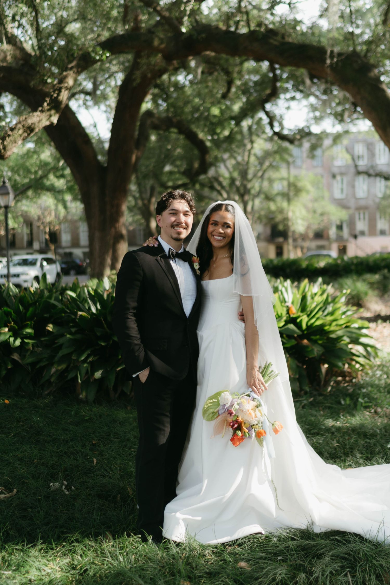 A wedding couple poses outdoors under a large oak tree in a garden setting with lush greenery.