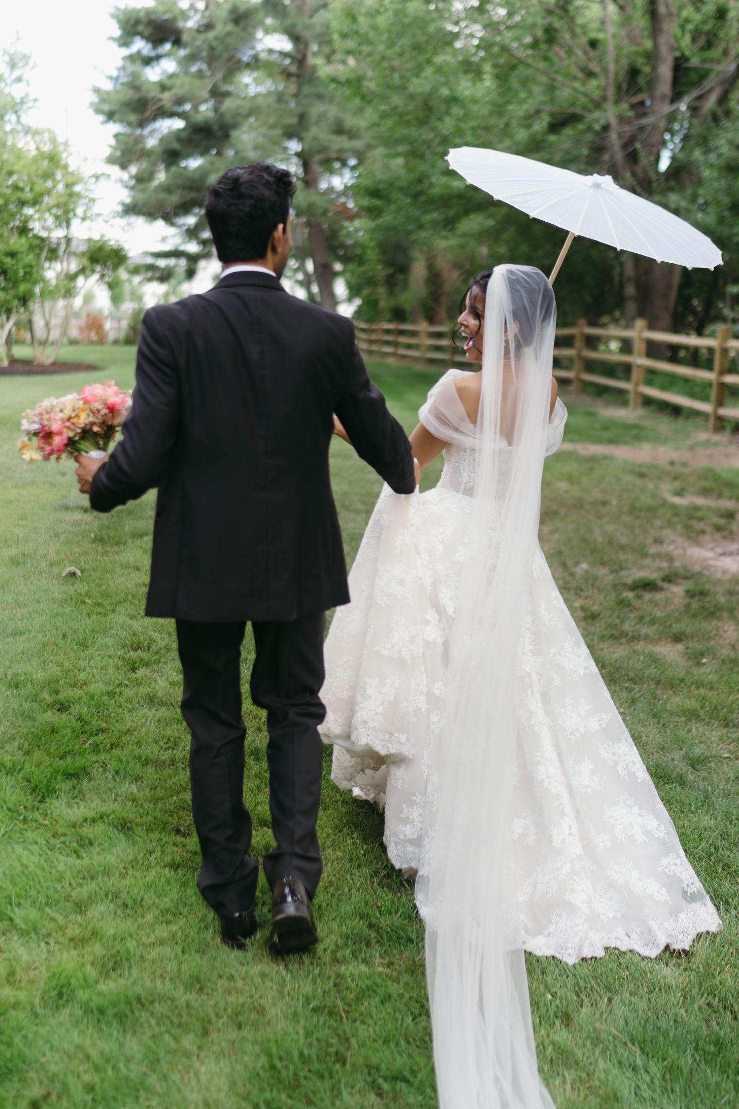 Bride and groom walk hand in hand on green grass, bride holding white parasol and lace veil trailing.