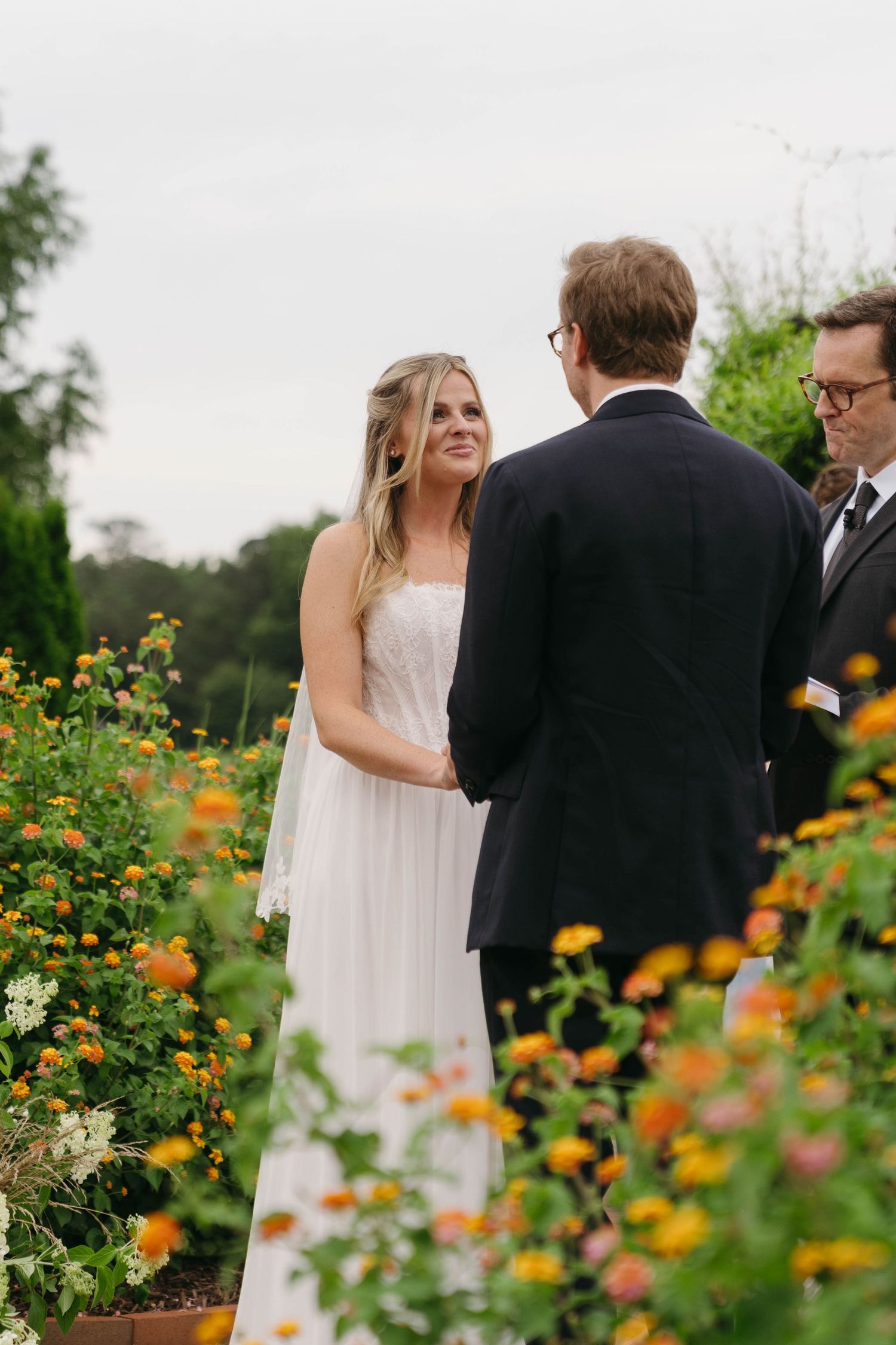Bride in white gown exchanges vows with groom outdoors surrounded by colorful garden flowers.