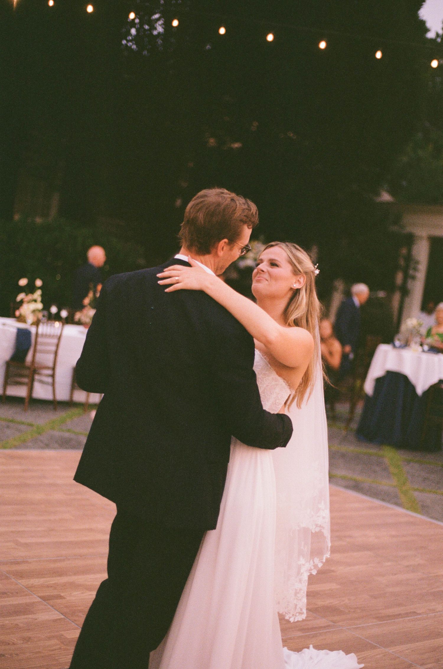Bride and groom share their first dance outdoors at evening wedding reception with string lights overhead.