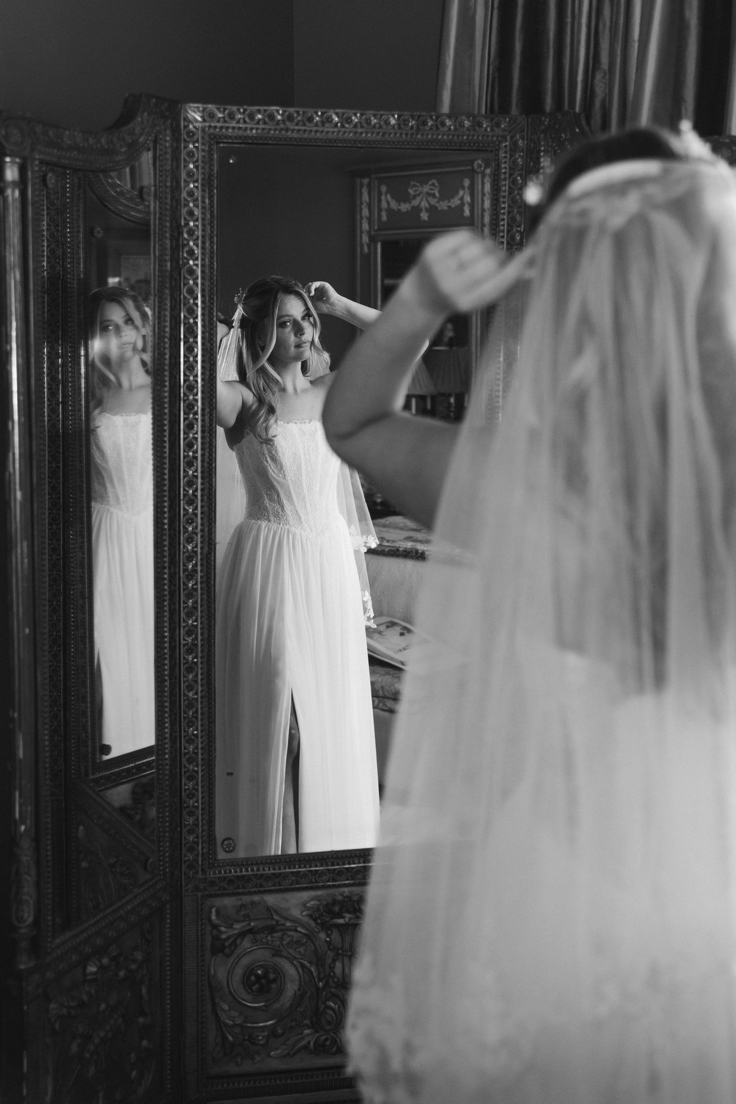 Black and white photo of bride adjusting her hair while viewing reflection in ornate vintage mirror.