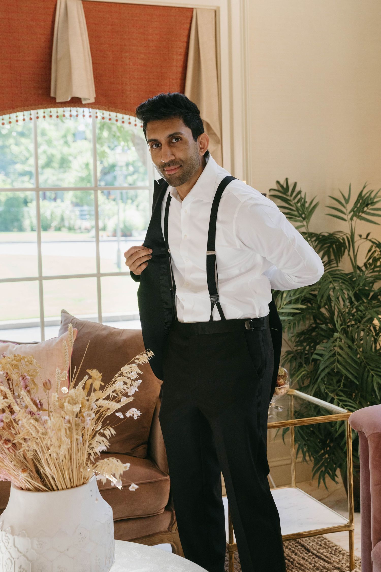 Groom in white shirt with suspenders and black vest getting dressed in elegant room before wedding.