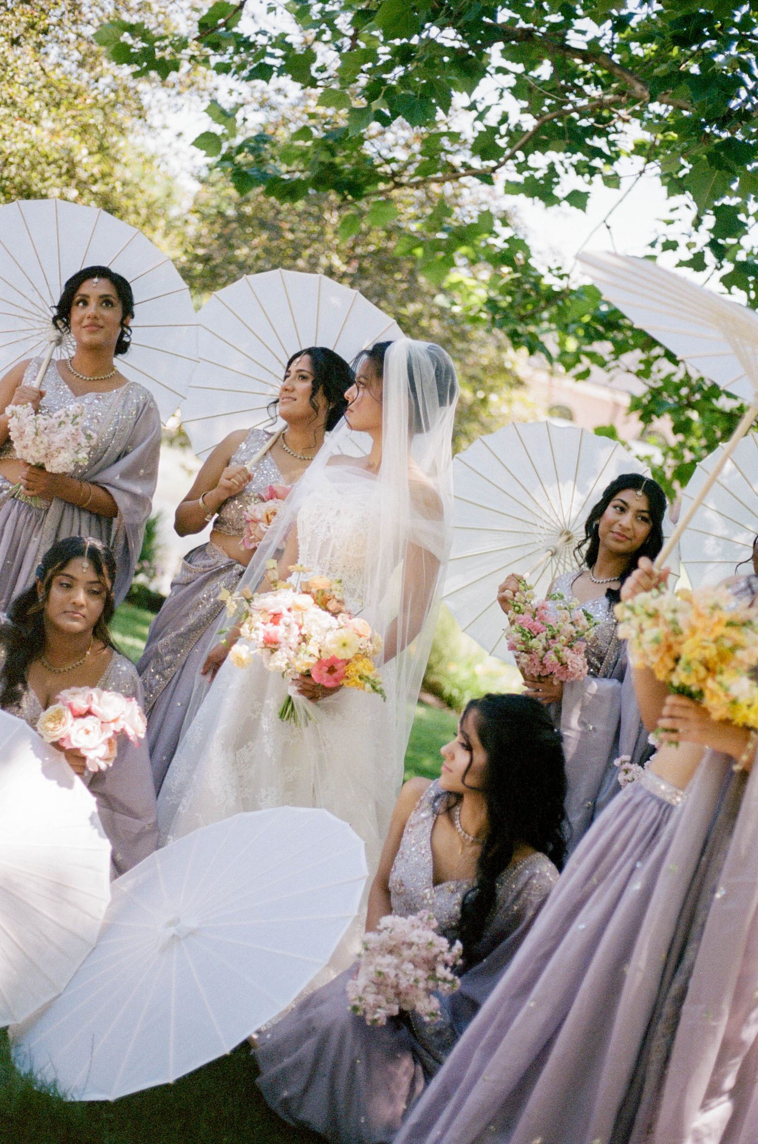 Bride surrounded by bridesmaids in lavender sarees holding white parasols and colorful bouquets outdoors.