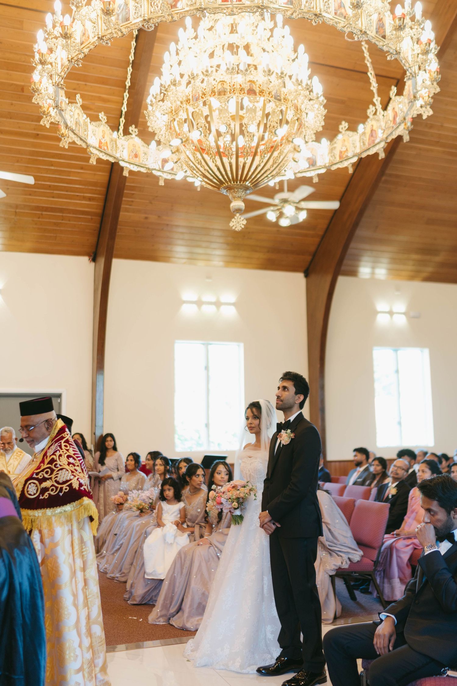 Bride and groom stand before an officiant during a wedding ceremony under a grand crystal chandelier.