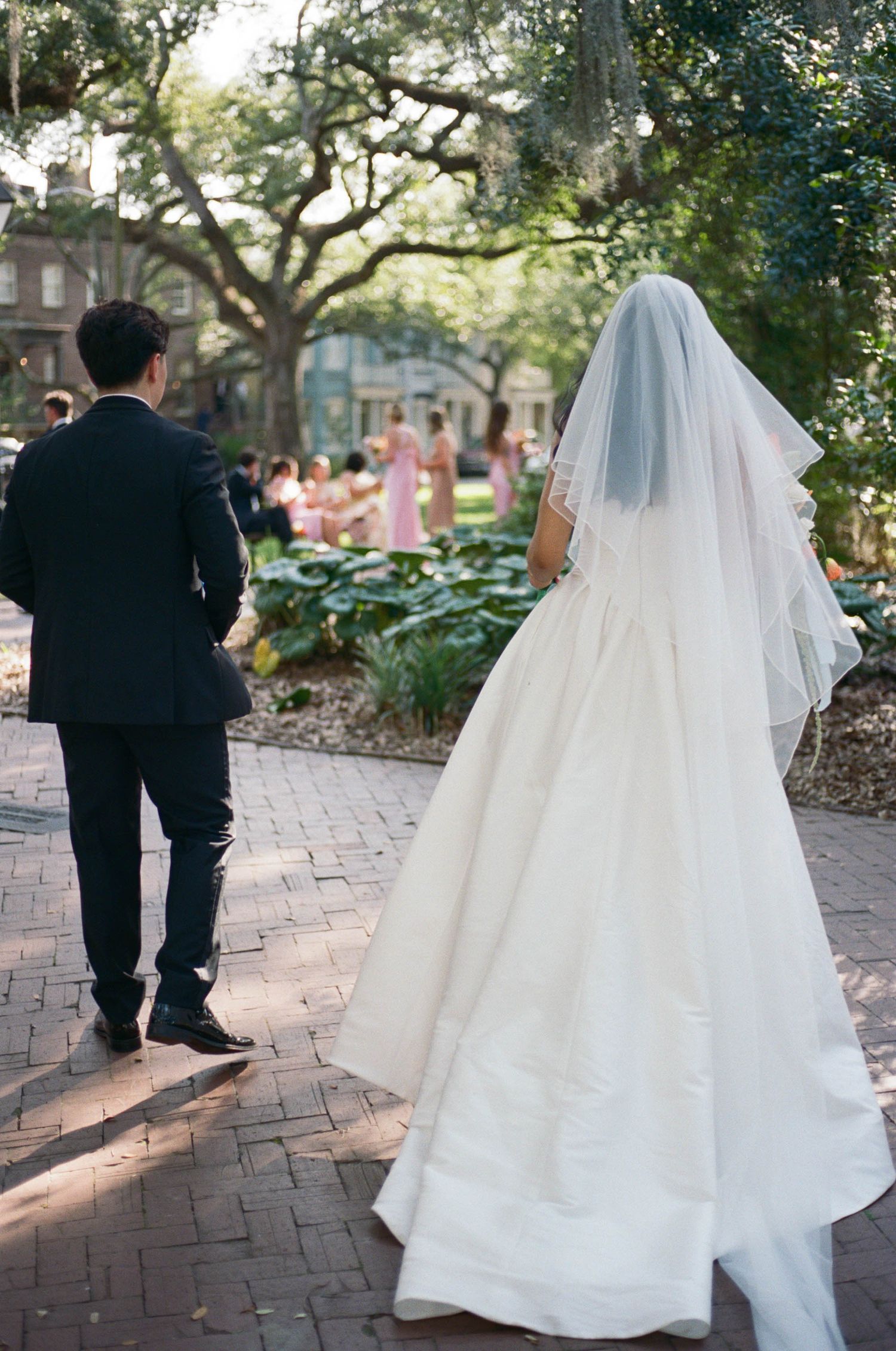 A bride in a flowing white gown and groom in black suit walk along a brick pathway under trees.