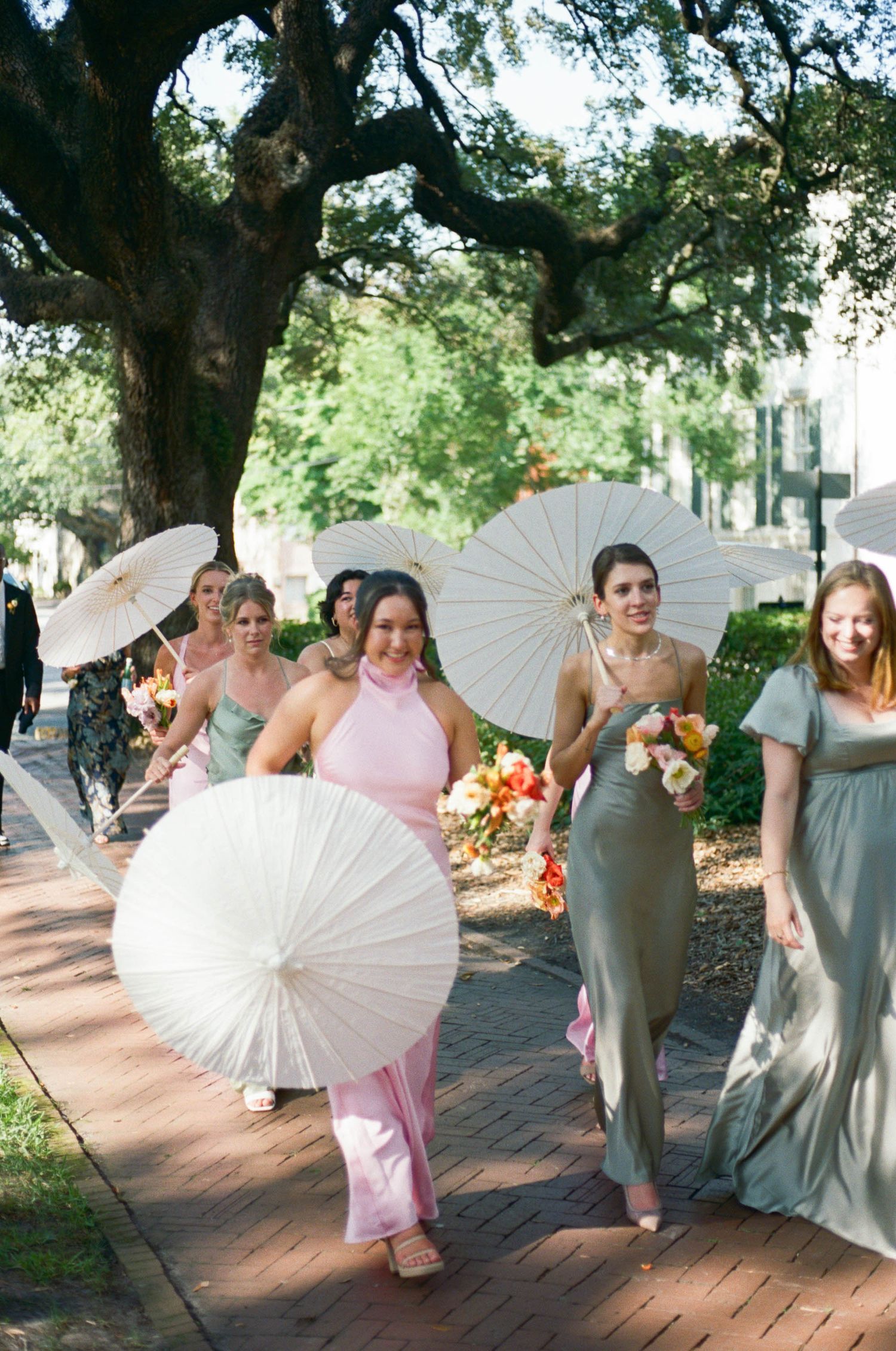 Bridesmaids in pastel dresses carry white parasols during outdoor wedding photos.
