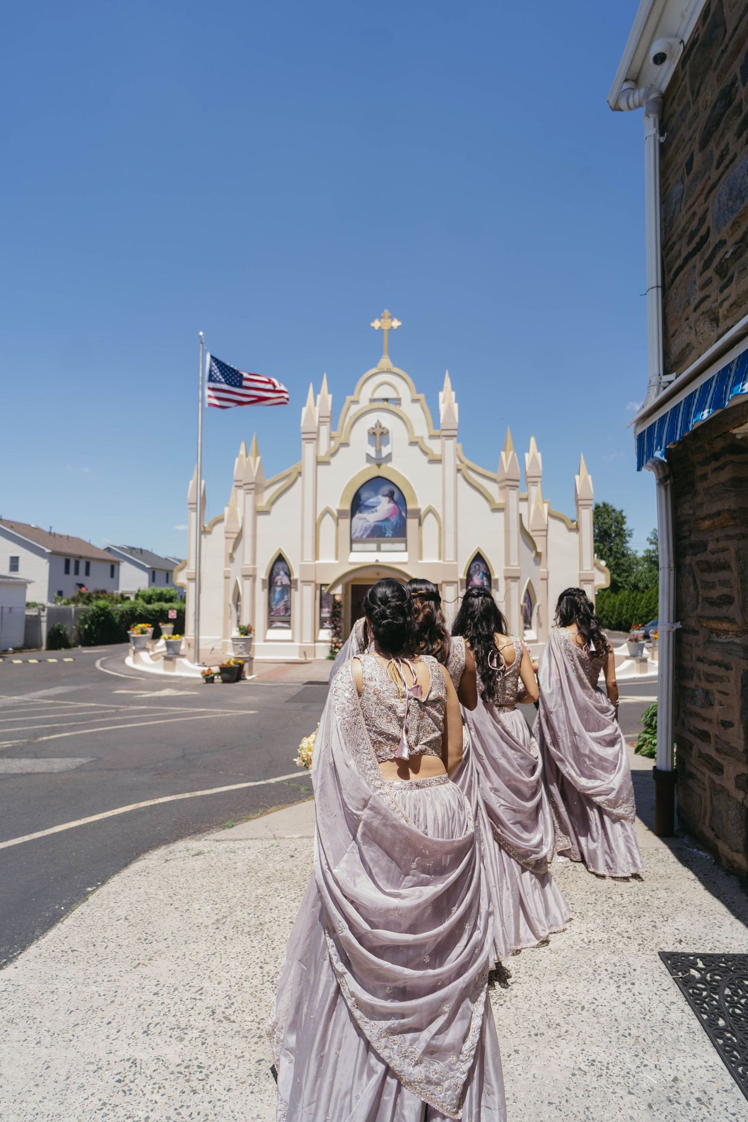 Bridesmaids in purple sarees walking toward ornate white Catholic church with American flag on clear day.