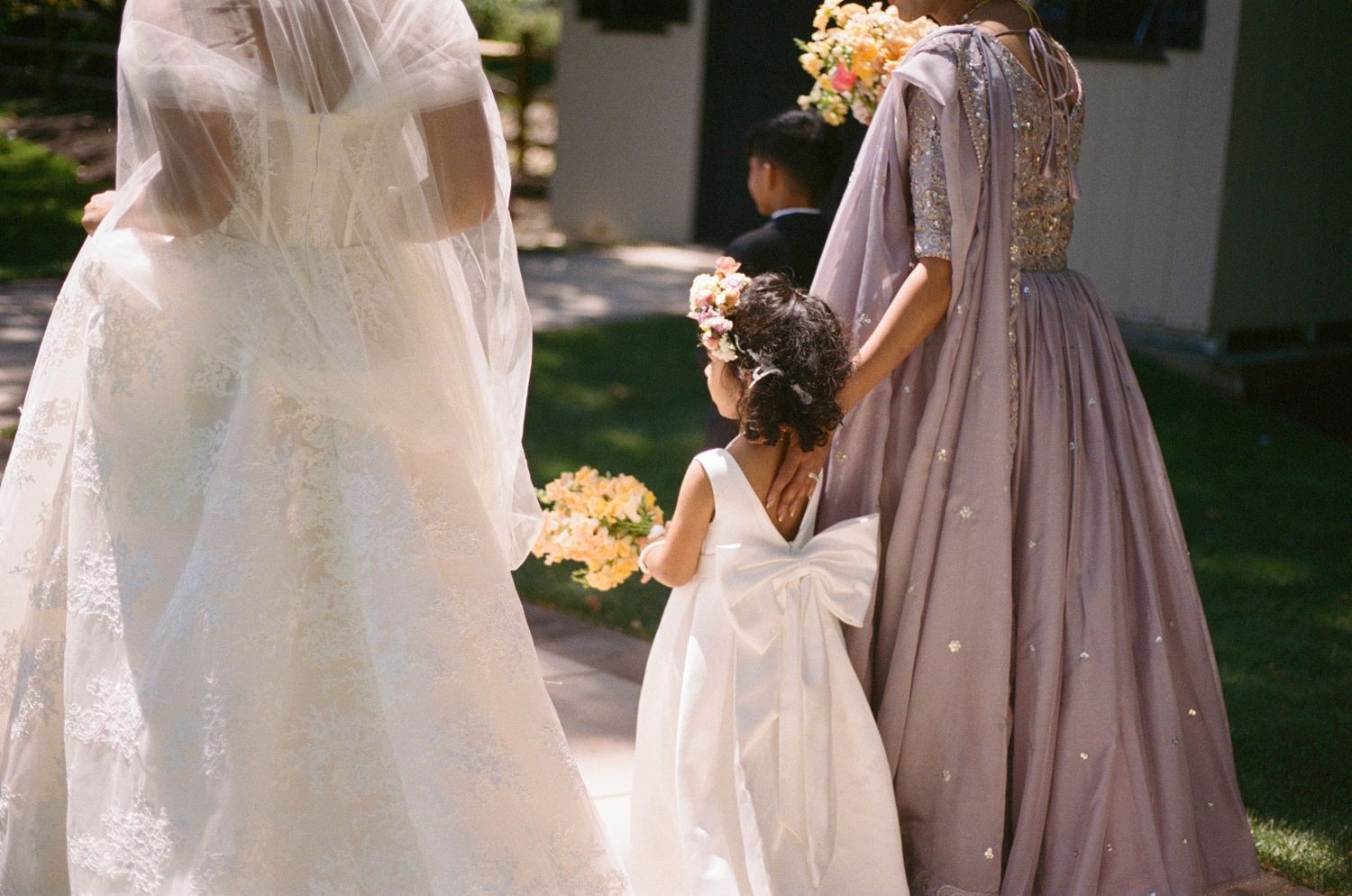 Bride in lace gown walks with flower girl and bridesmaid in mauve dress holding bouquets.