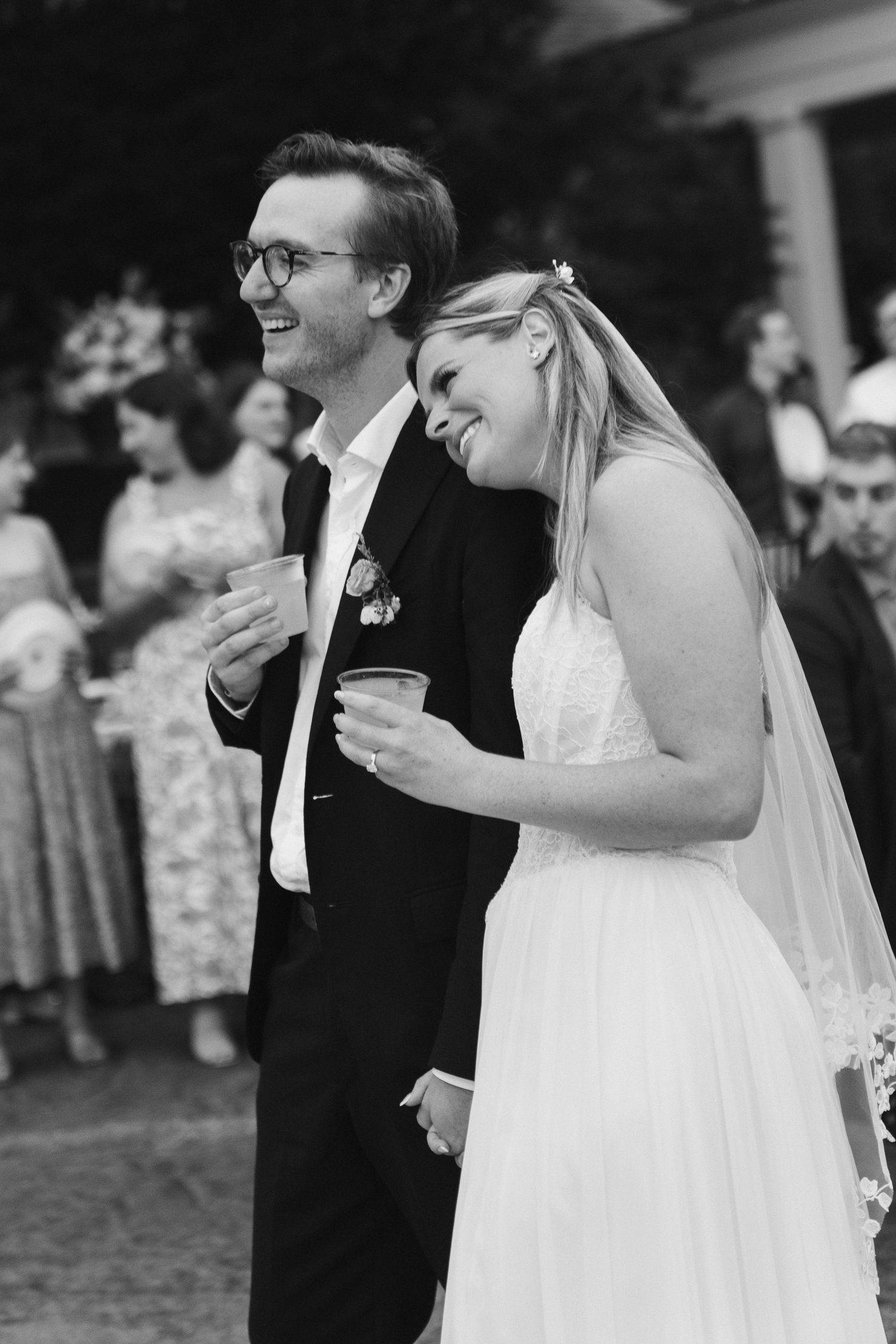 Black and white photo of smiling bride and groom holding drinks and laughing together at outdoor wedding reception.