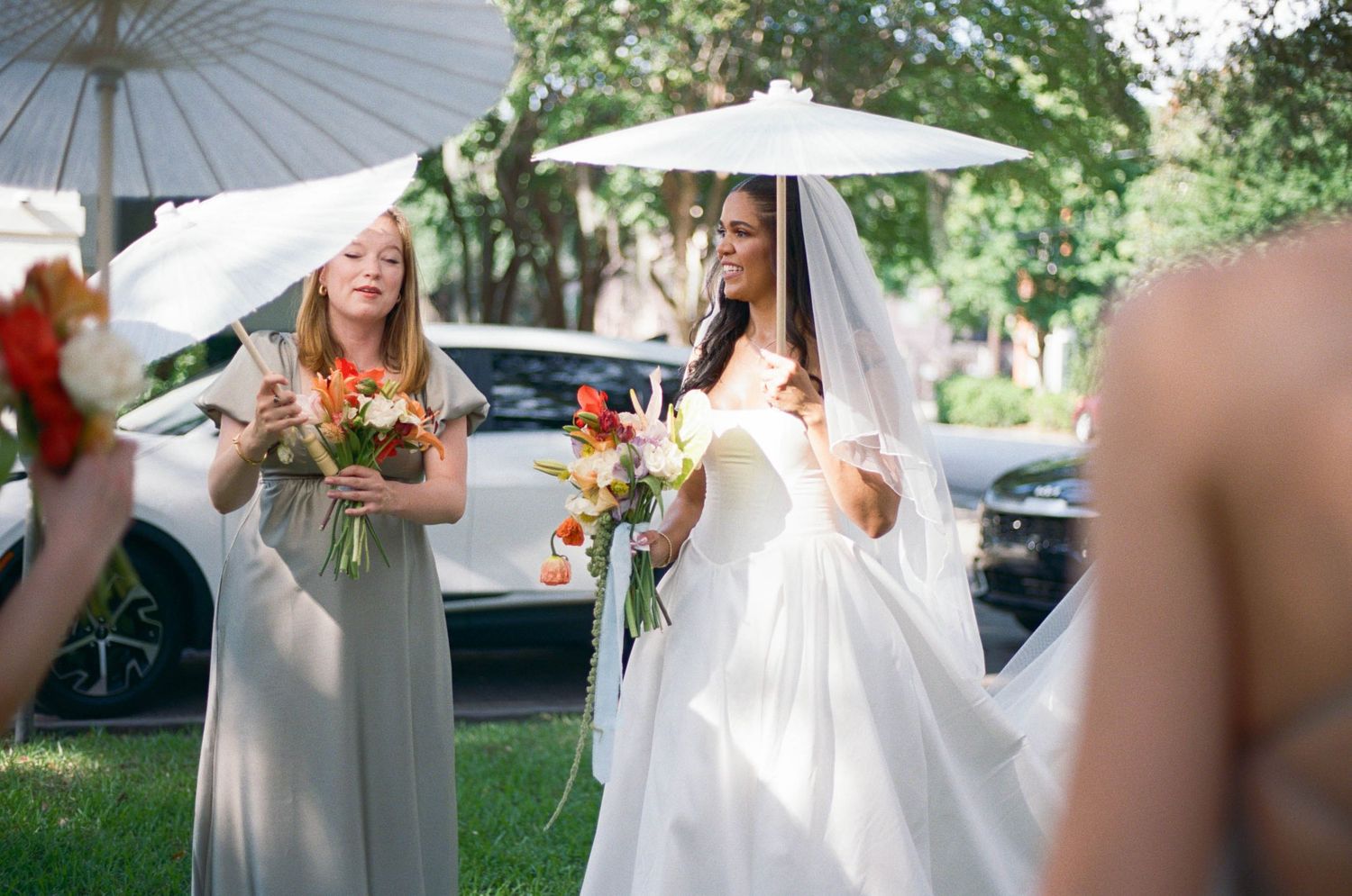 Wedding party members hold white parasols while walking outdoors.