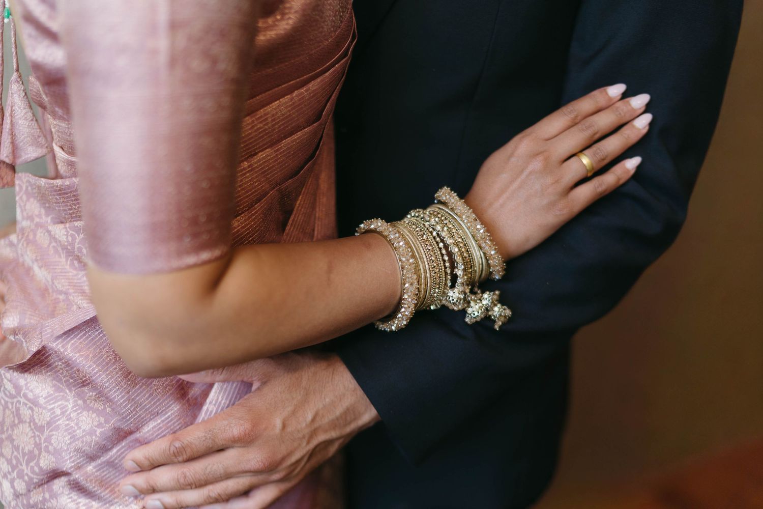 Close-up of bride wearing gold bangles and bracelets in a pink sari embracing groom in black suit.