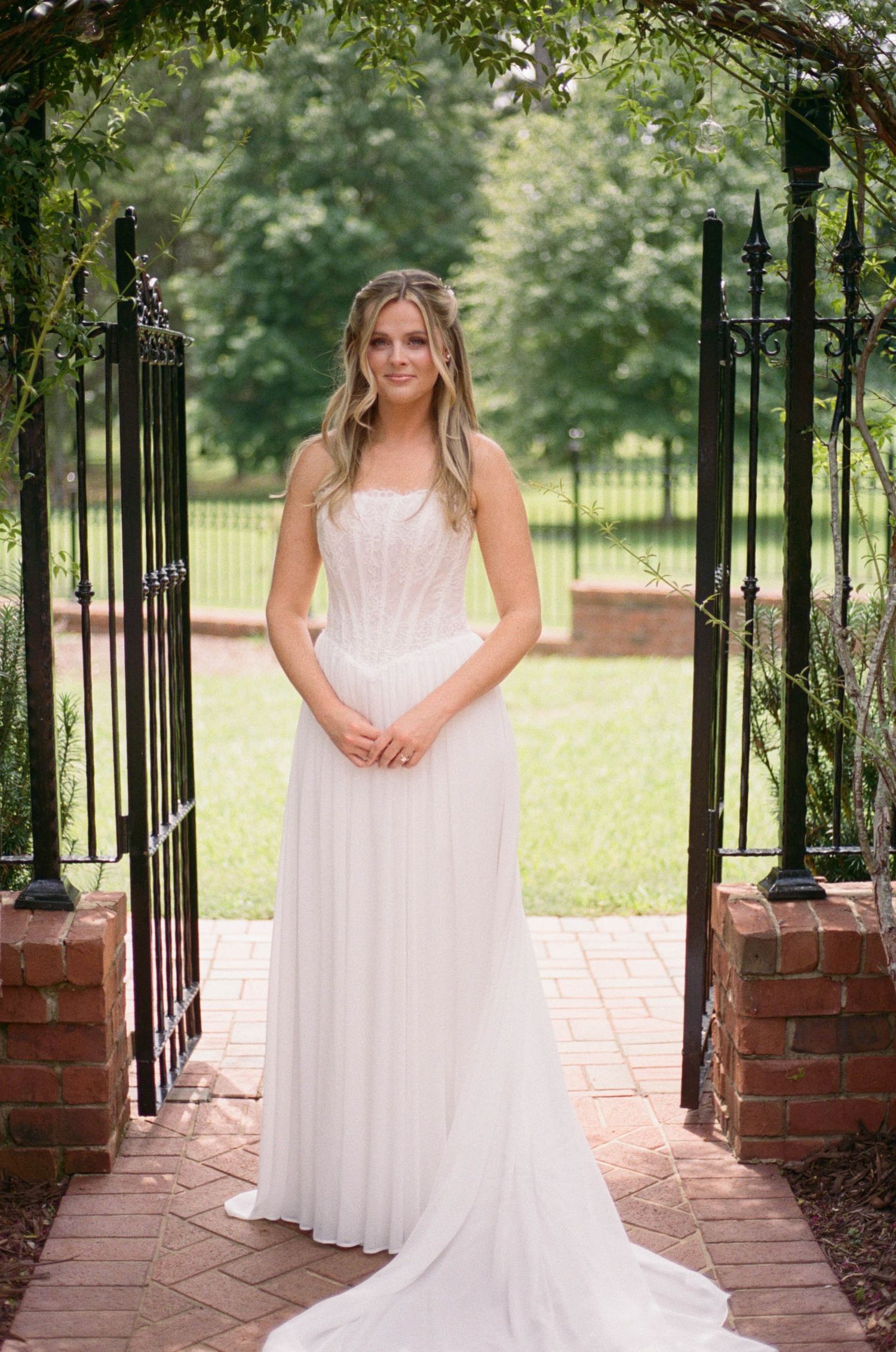 Bride in strapless white gown stands gracefully between ornate iron garden gates.