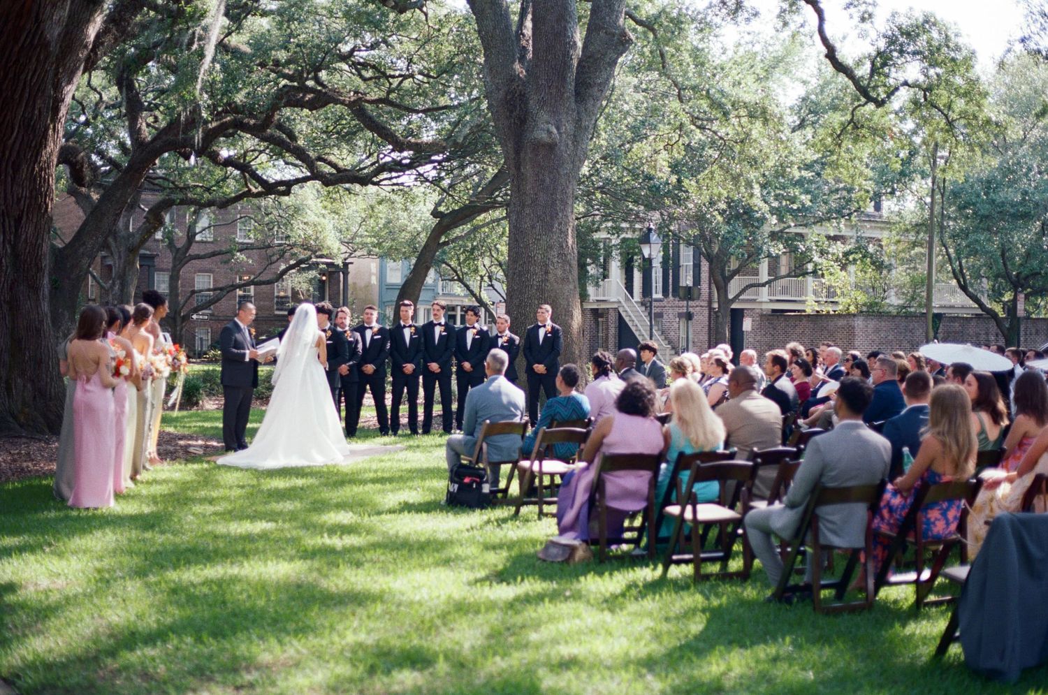 An outdoor wedding ceremony under large oak trees with guests seated on folding chairs.