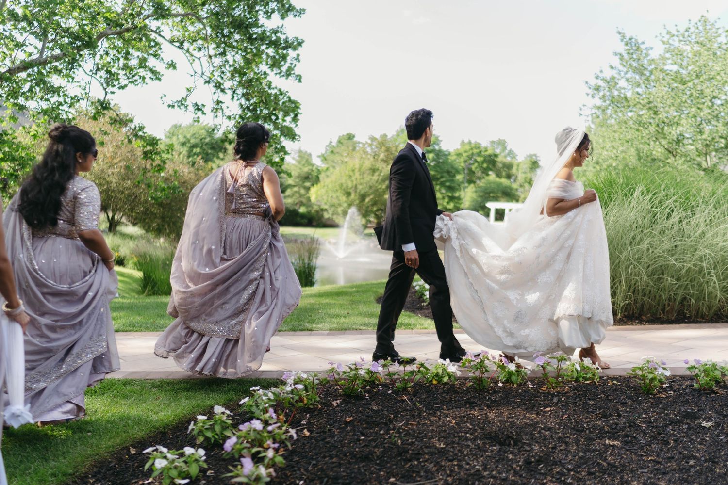 Bride in white gown walks with groom and bridesmaids in lavender dresses through a lush garden.