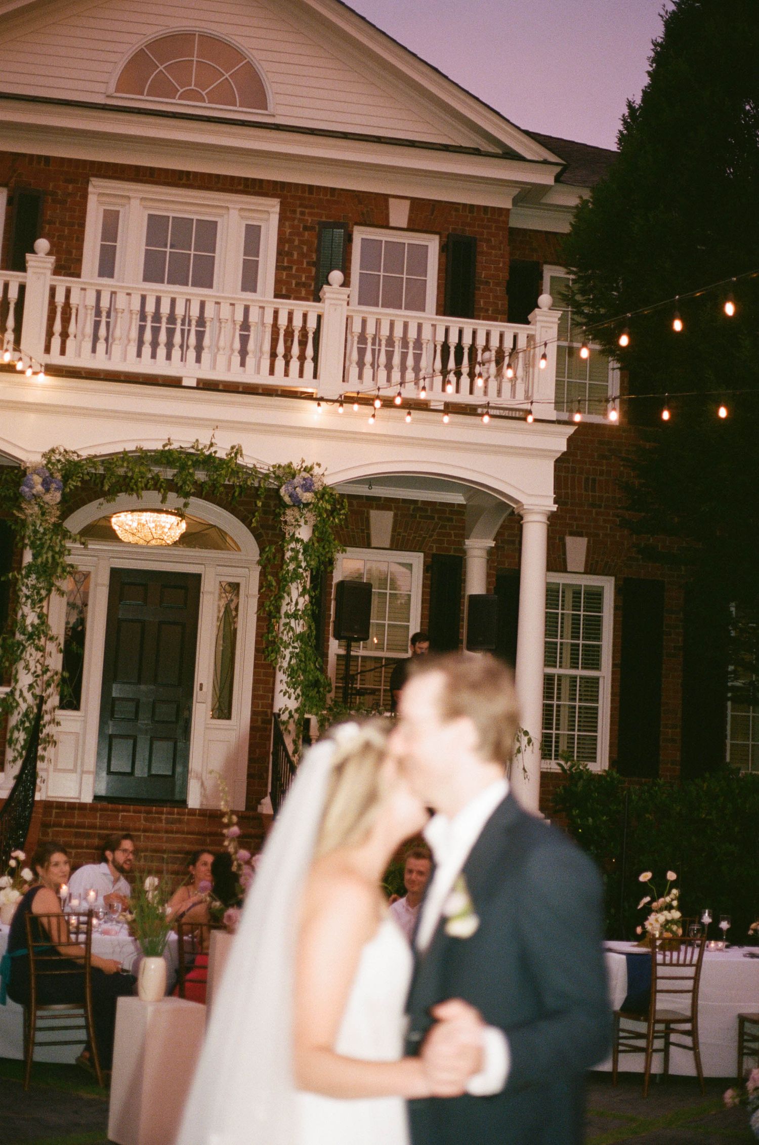 Bride and groom share first dance kiss at twilight outside elegant brick mansion with string lights.