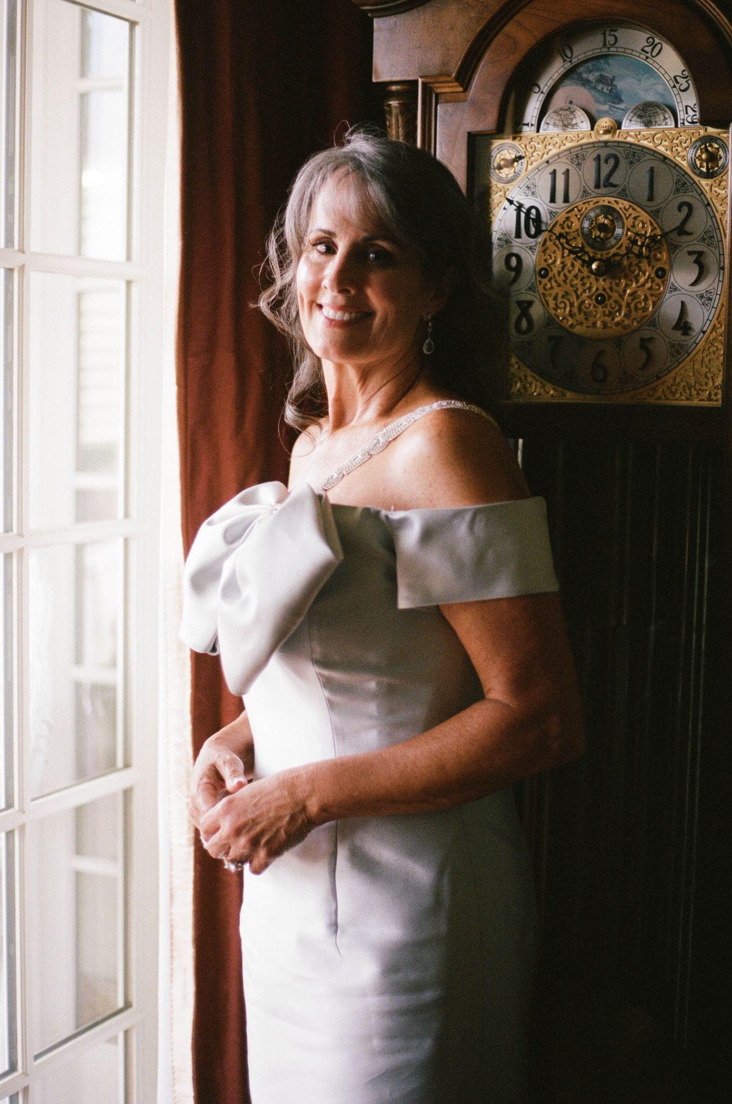 Elegant woman in off-shoulder white gown smiling beside a grandfather clock near a window.