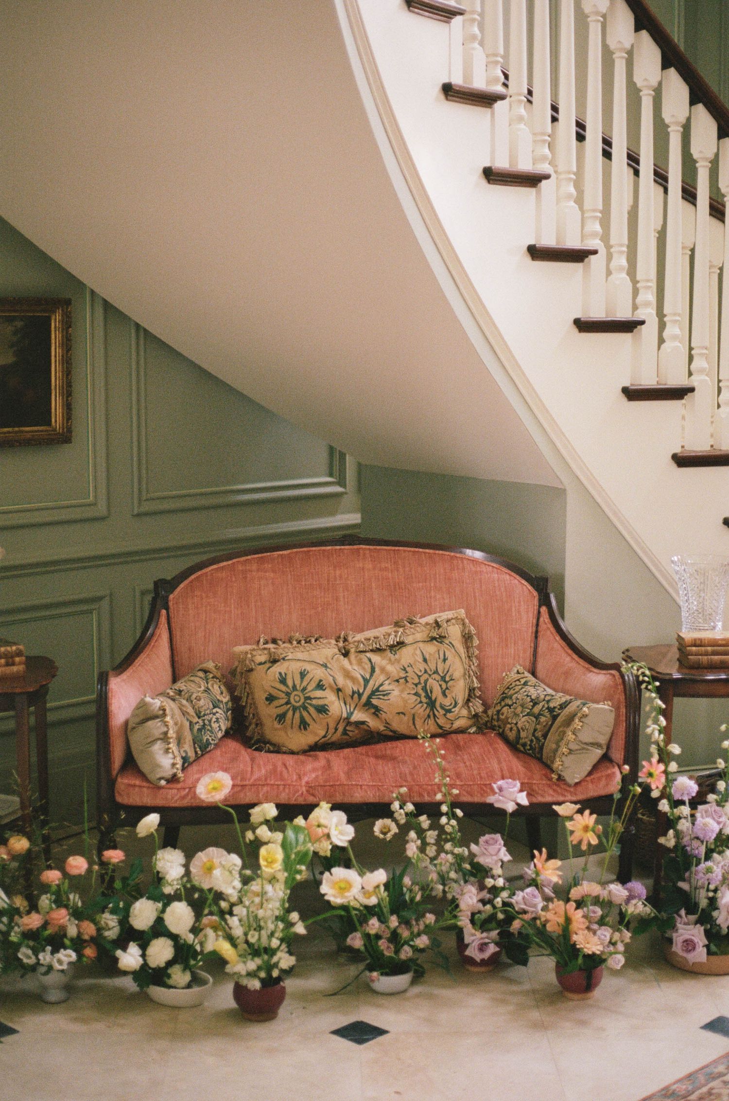 Elegant pink antique sofa beneath curved staircase surrounded by colorful floral arrangements in green room.