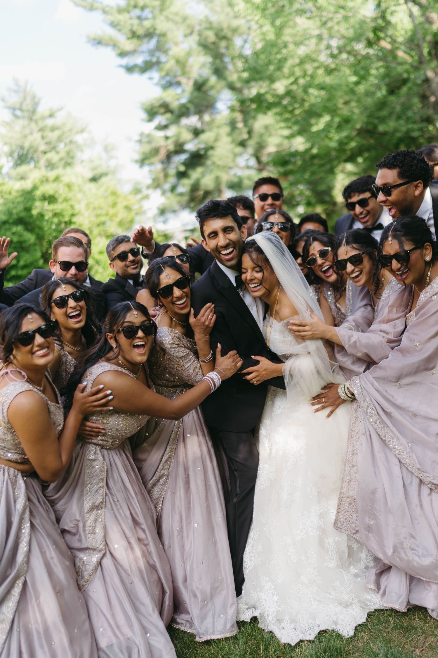 Bride and groom surrounded by laughing wedding party in lavender sarees and sunglasses outdoors.