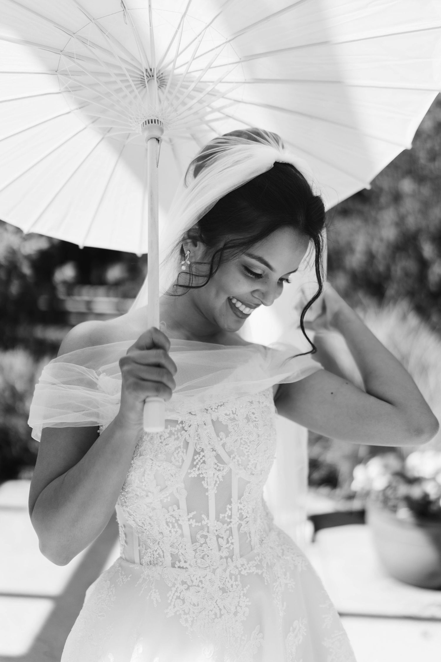Smiling bride in lace wedding dress holding white parasol umbrella, black and white bridal portrait.