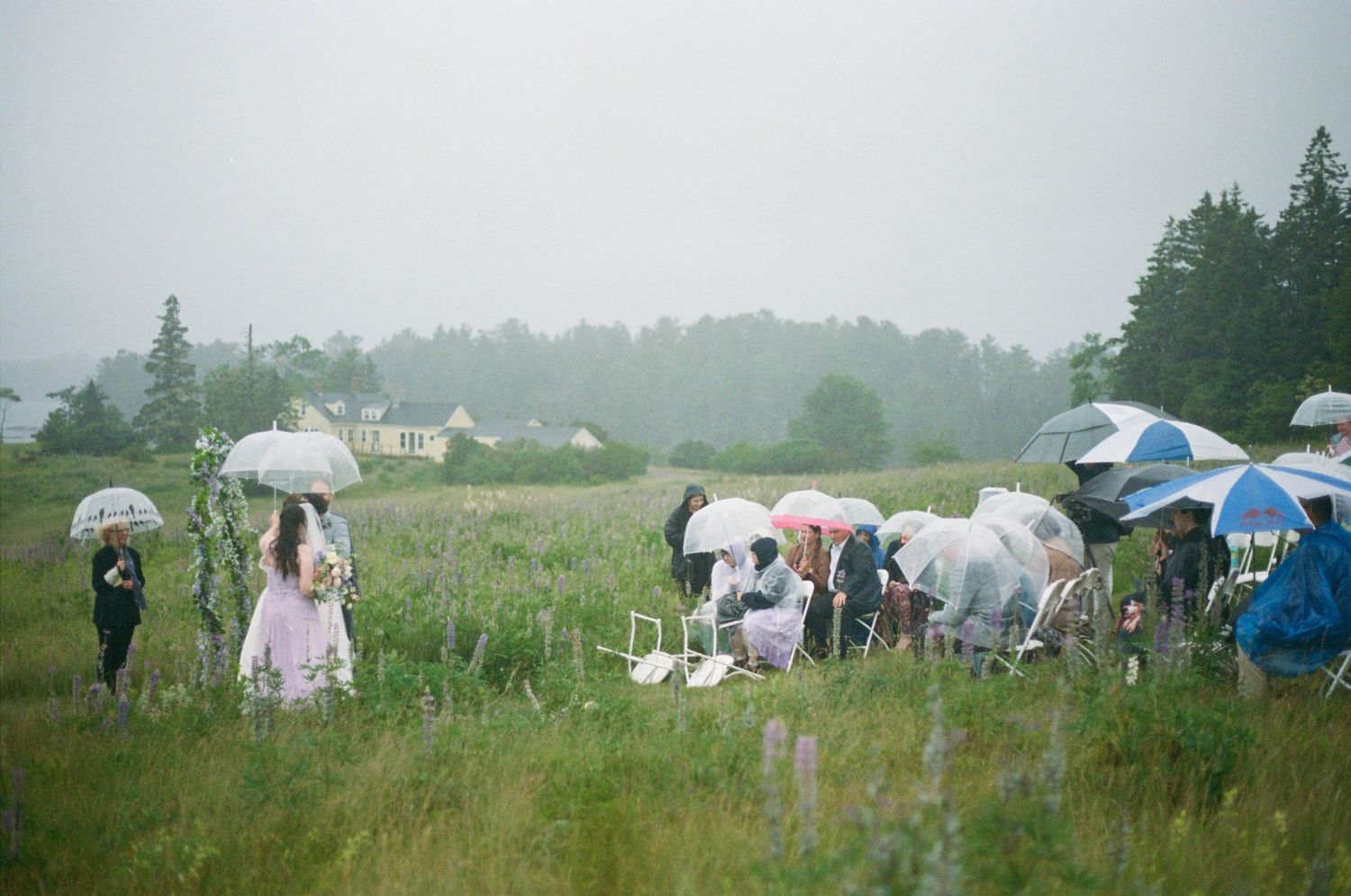 Wedding guests gather in a misty field with white umbrellas and tents during an outdoor ceremony.