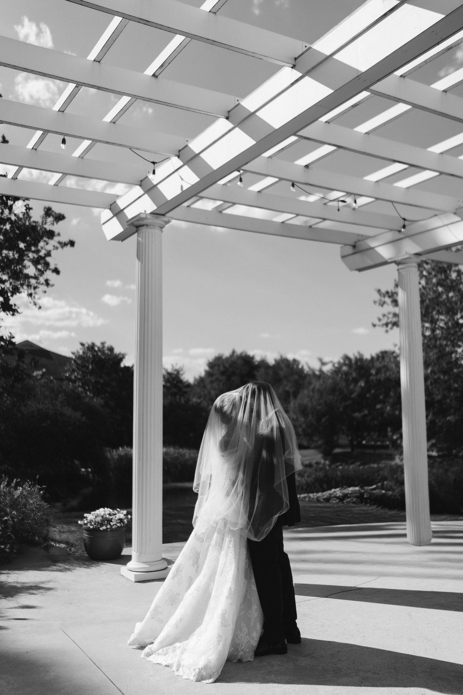 Black and white photo of bride and groom kissing under elegant white pergola at outdoor wedding venue.
