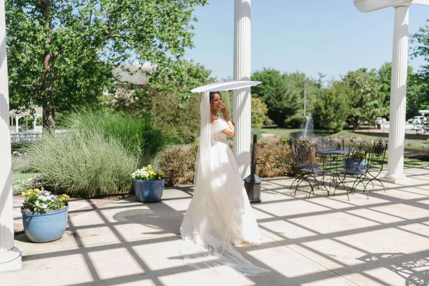 Bride in white lace gown and veil holding bouquet under pergola in sunny outdoor garden setting.