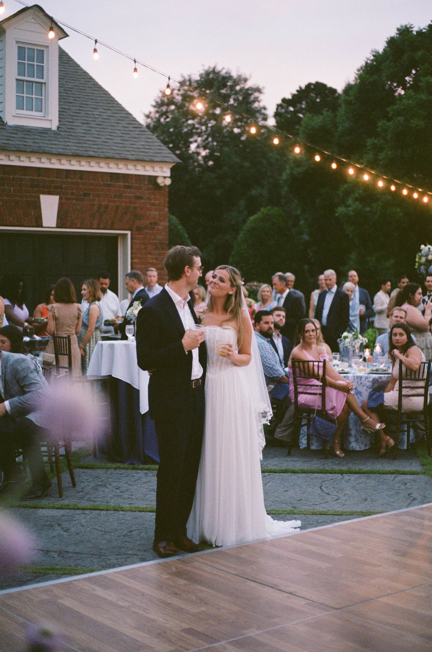 Bride and groom share first dance at elegant outdoor wedding reception with string lights and seated guests.