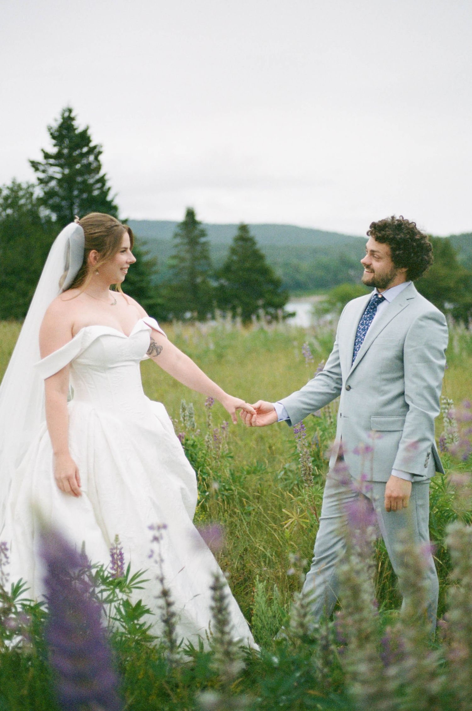 Couple walking hand in hand through a meadow of purple wildflowers on their wedding day.