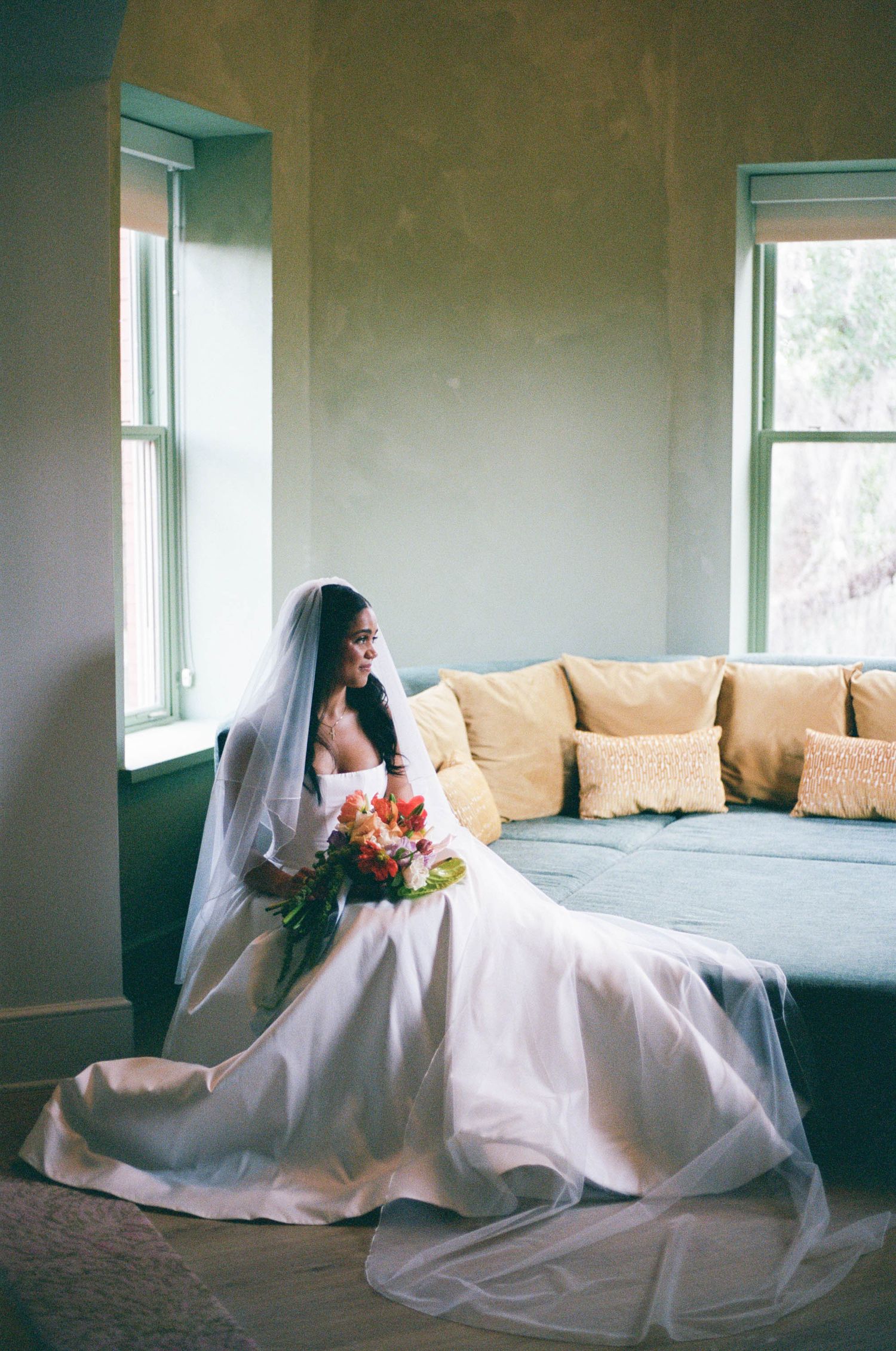 A bride in white dress sits gracefully on a couch near windows in a minimalist room.