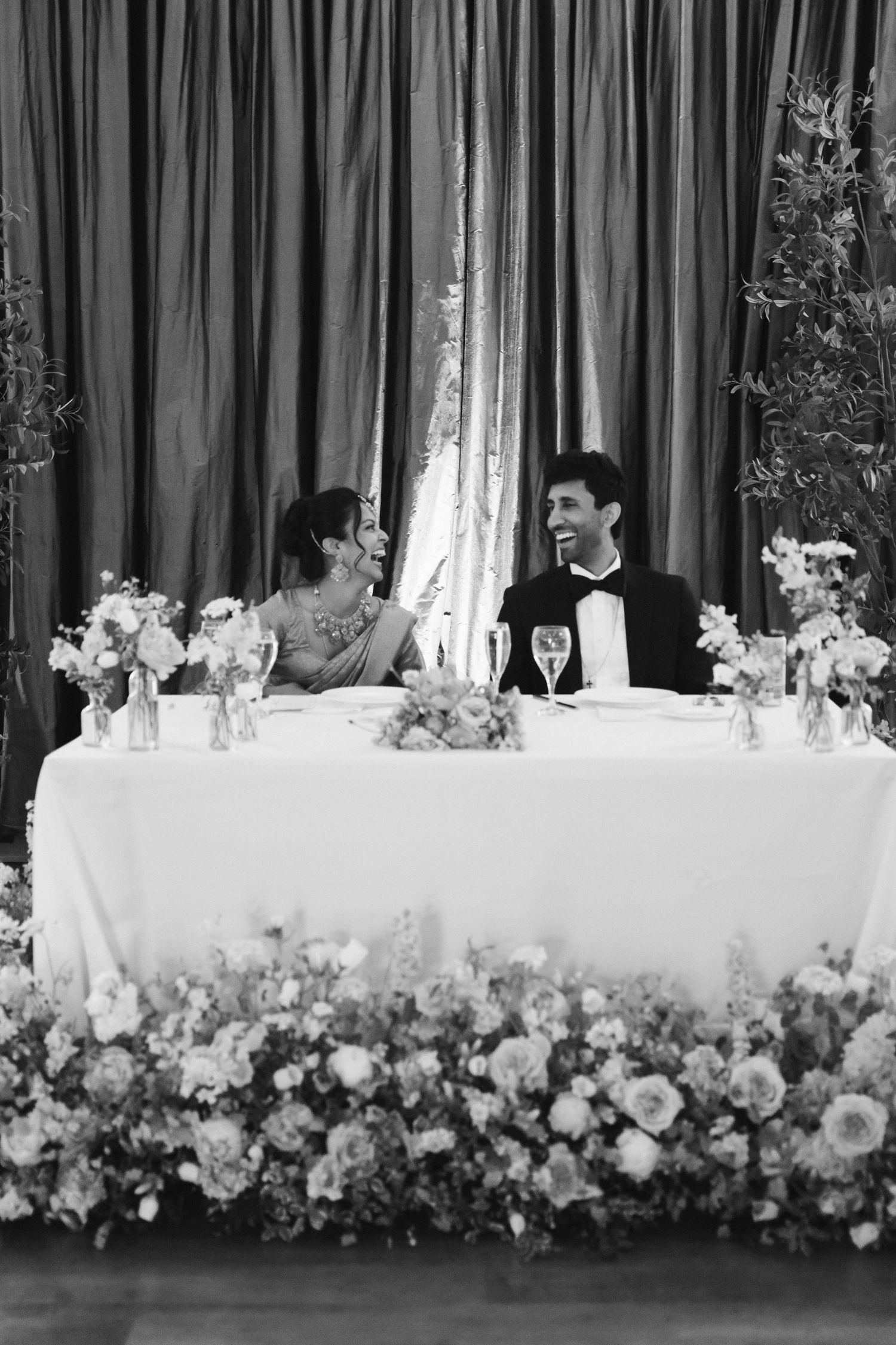 Bride and groom share a joyful moment at their elegantly decorated sweetheart table with floral arrangements.