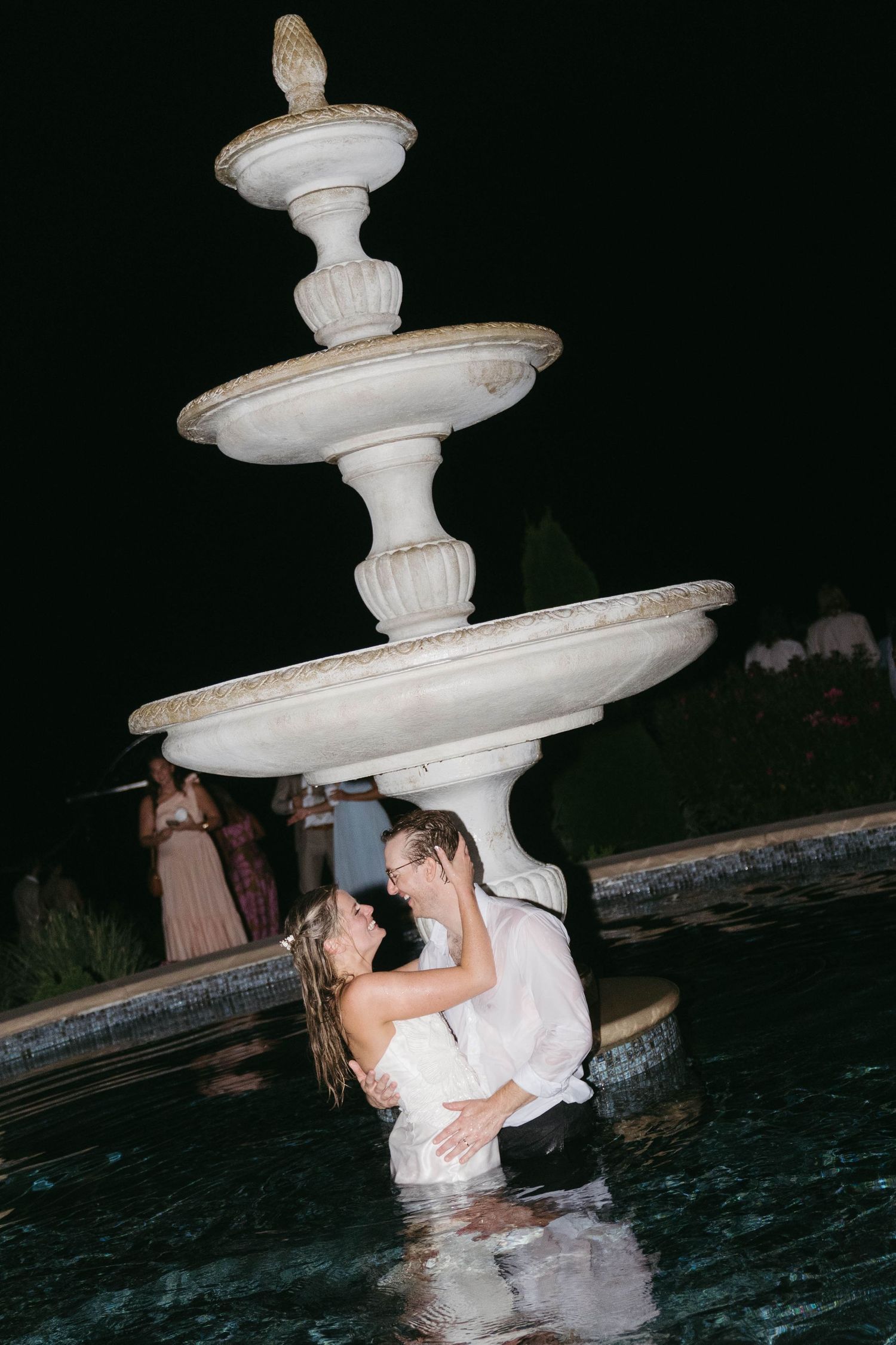 Bride and groom embracing in a fountain at night during their outdoor wedding celebration.